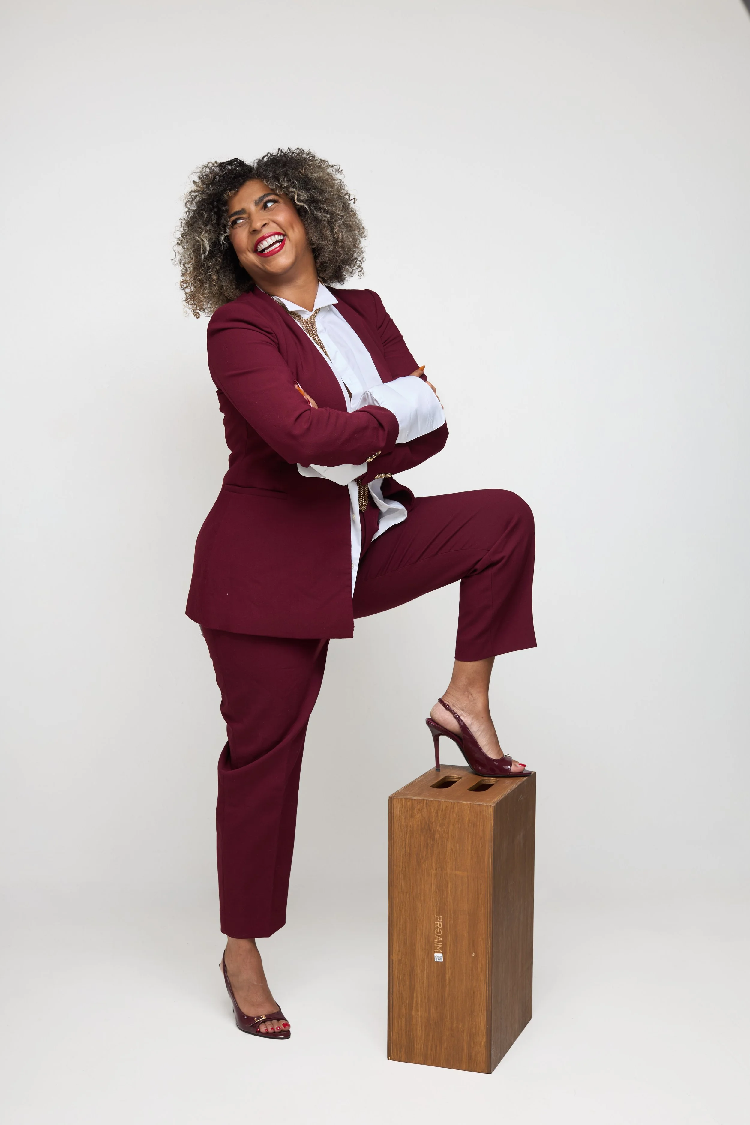 Femme souriante aux cheveux bouclés gris, en tailleur bordeaux et chemisier blanc, posant avec un pied sur un cube en bois.