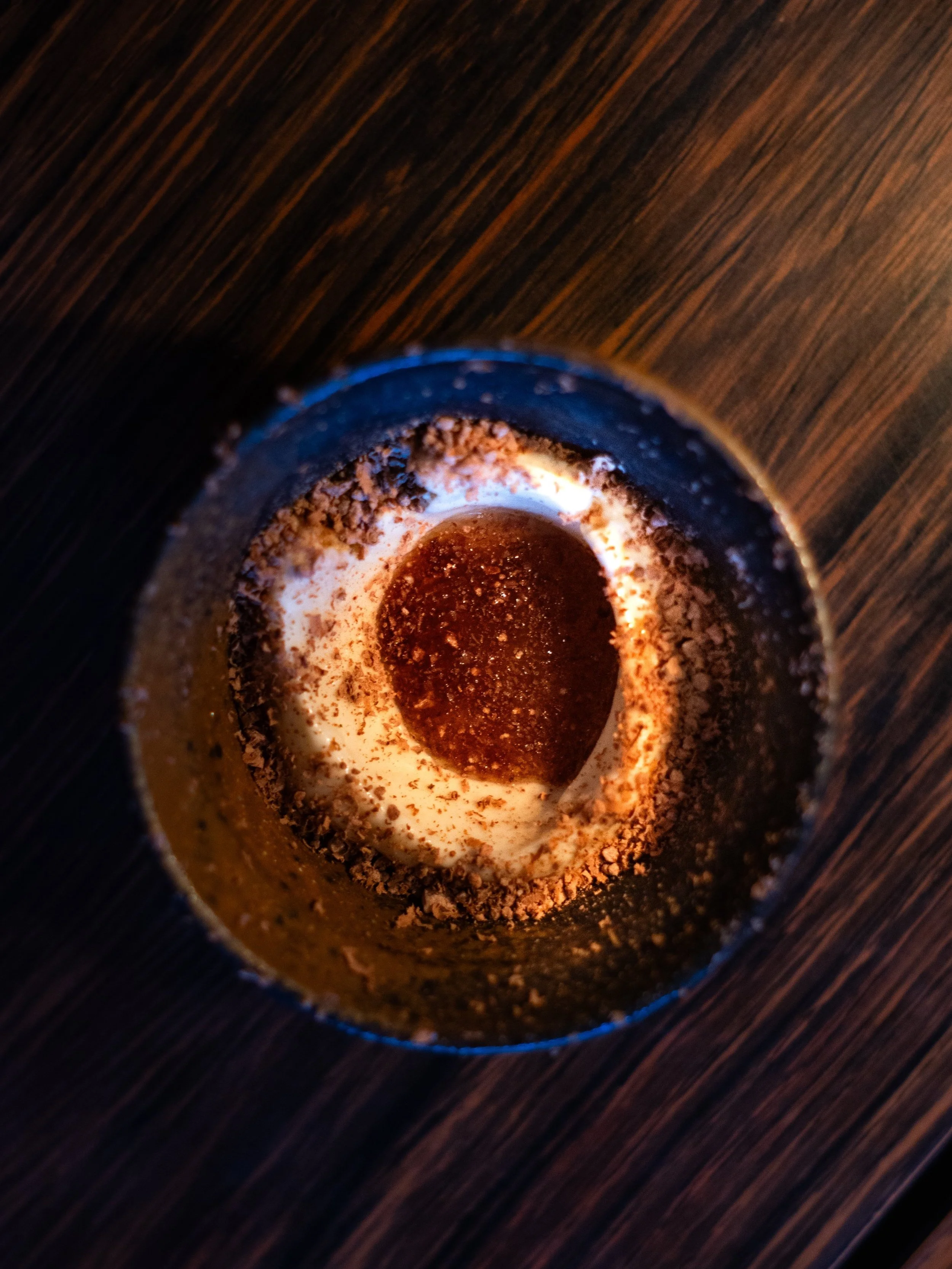 Top view of a dessert in a glass cup topped with cocoa powder, whipped cream, and a chocolate heart, on a wooden surface.