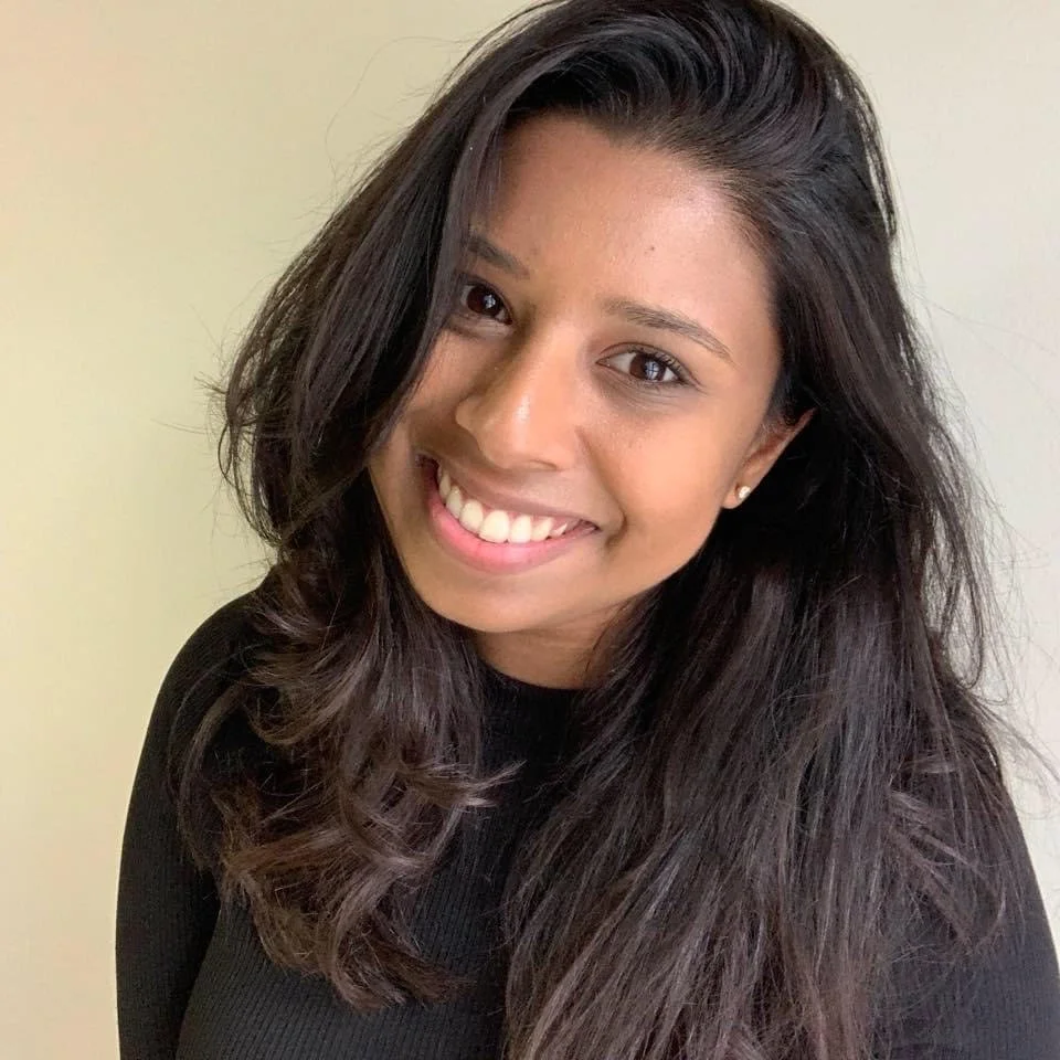 A young woman with long, dark, wavy hair smiling at the camera against a plain background.