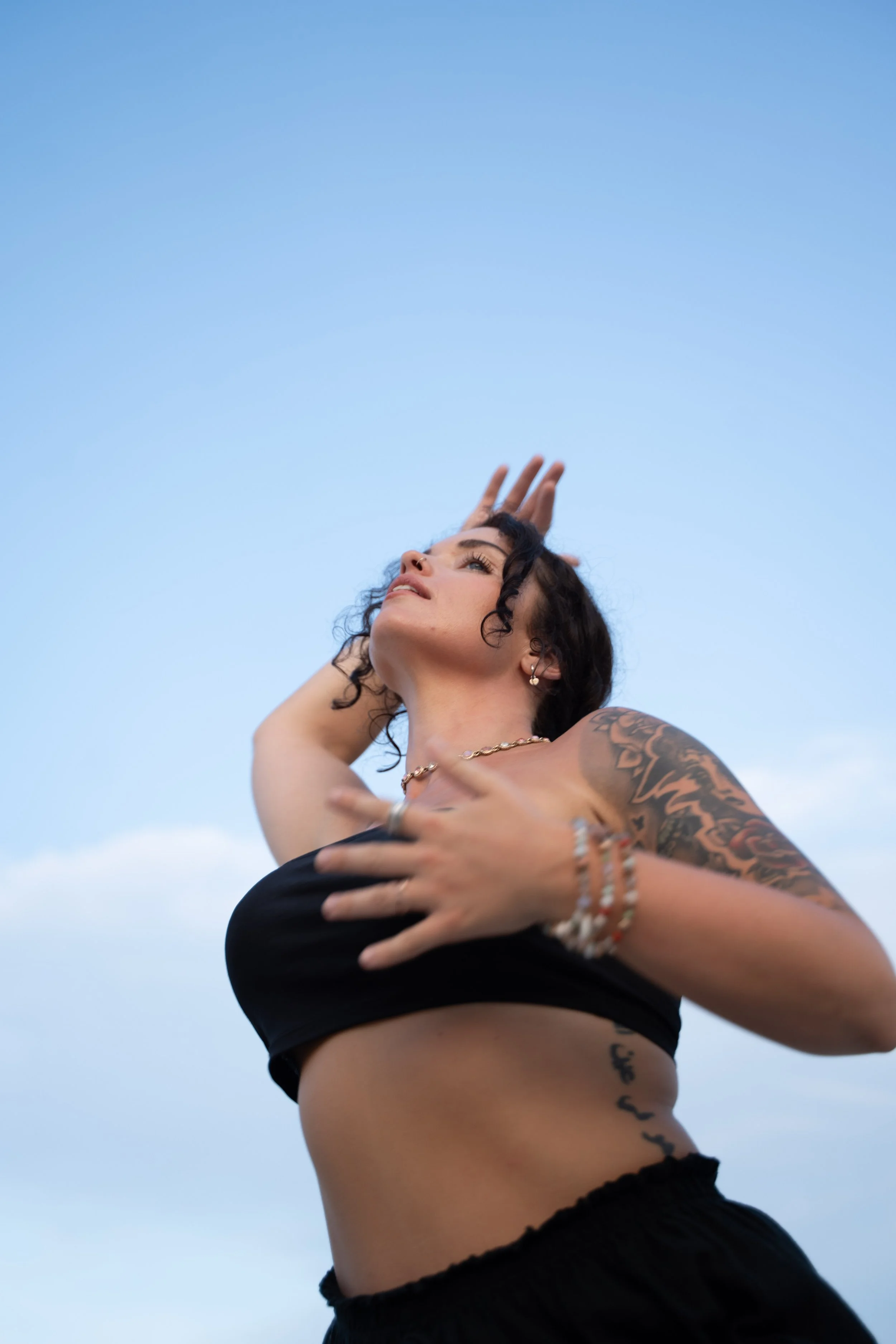 A woman with tattoos and jewelry poses outdoors against a clear blue sky, wearing a black crop top and black pants, with her hand touching her head.