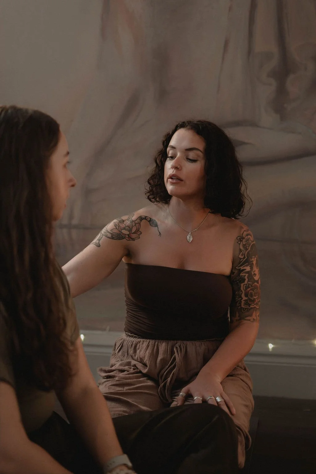 Two women sitting and talking indoors, one with dark curly hair, tattoos on arms, and wearing a black strapless top and brown pants, the other with long brown hair, wearing a dark shirt. The background appears to be a large painted or printed mural.