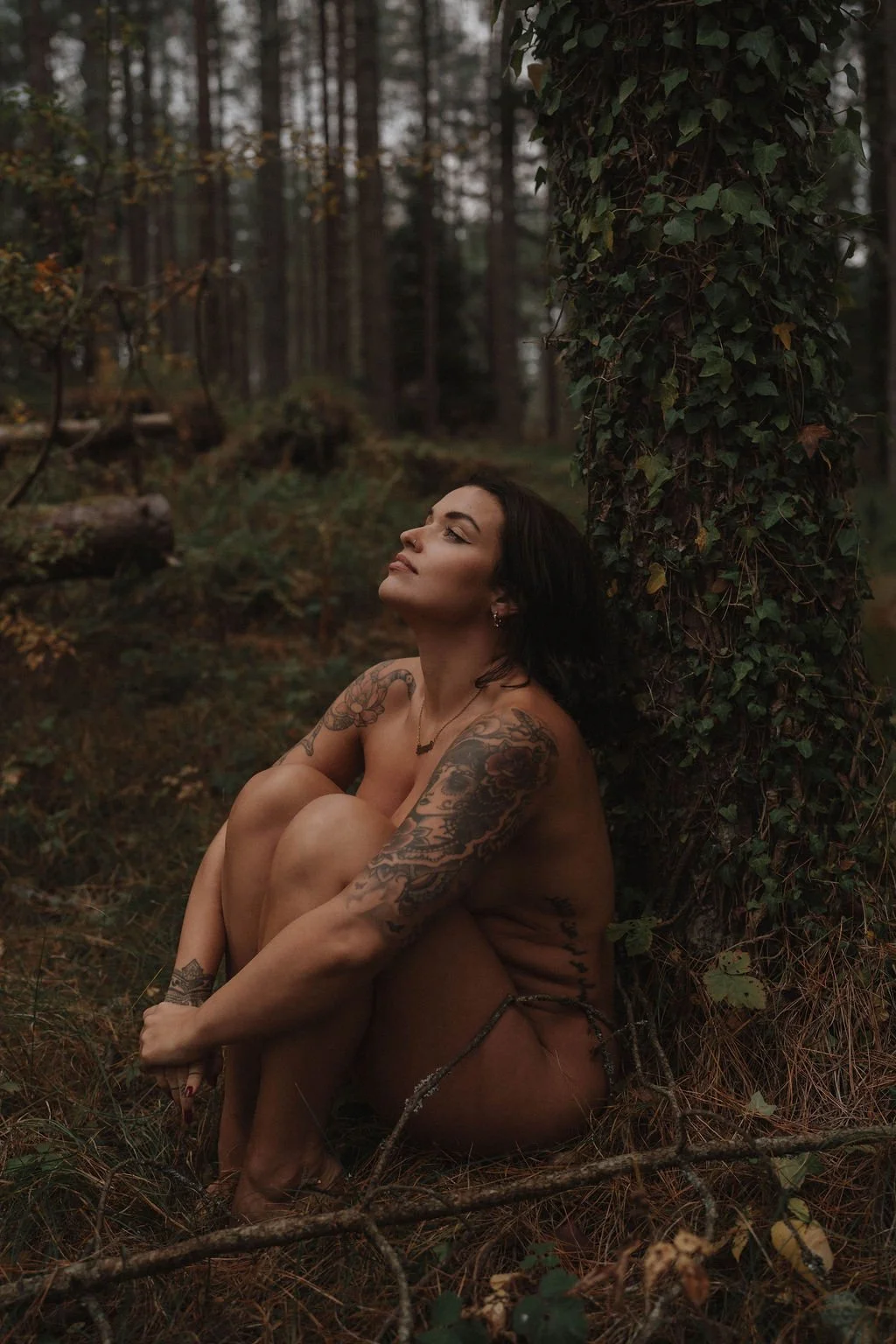 A woman with tattoos sitting outdoors in a forest, leaning against a tree covered in ivy.