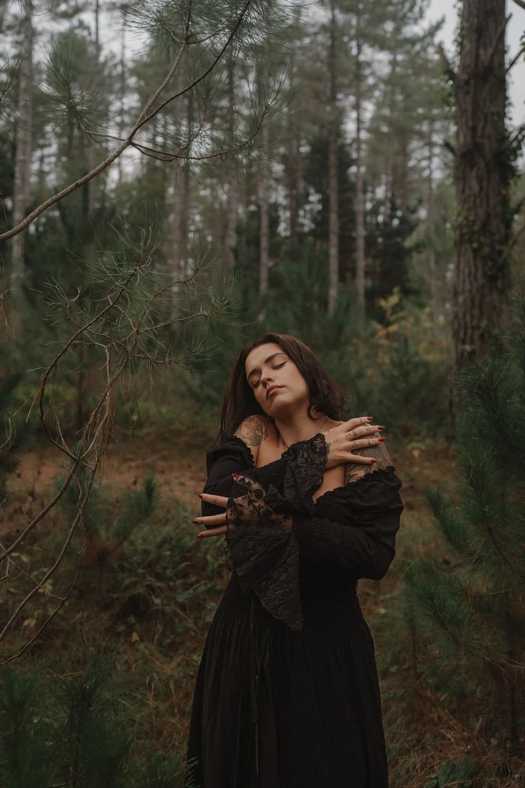 A woman with dark hair and tattoos, dressed in black with lace details, stands in a forest with her eyes closed, arms crossed over her chest.