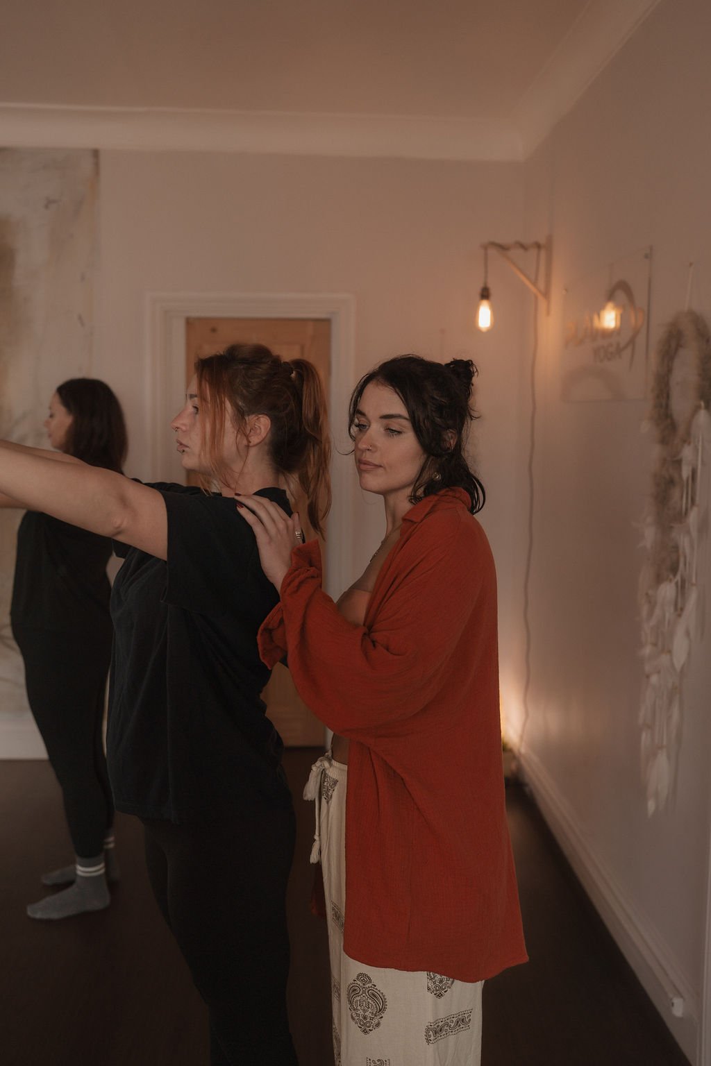 Three women stand in a dimly lit room, with one woman with red hair and a black shirt standing in front with arms extended, and a woman with black hair and a rust-colored cardigan gently touching her shoulder, both appearing to be engaged in a dance or yoga practice.