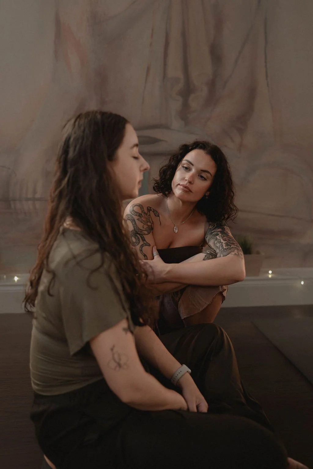 Two women sitting on a dark sofa, one with long dark hair in profile, the other with curly dark hair and tattoos on her arms, facing and looking at the woman with her arms crossed, in a dimly lit room with a blurred mural or painting in the background.