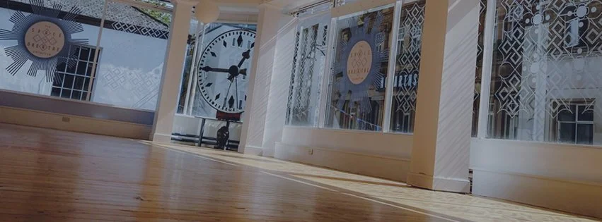Indoor space with large walls of windows, a wooden floor, and prominent clock faces visible through the windows.