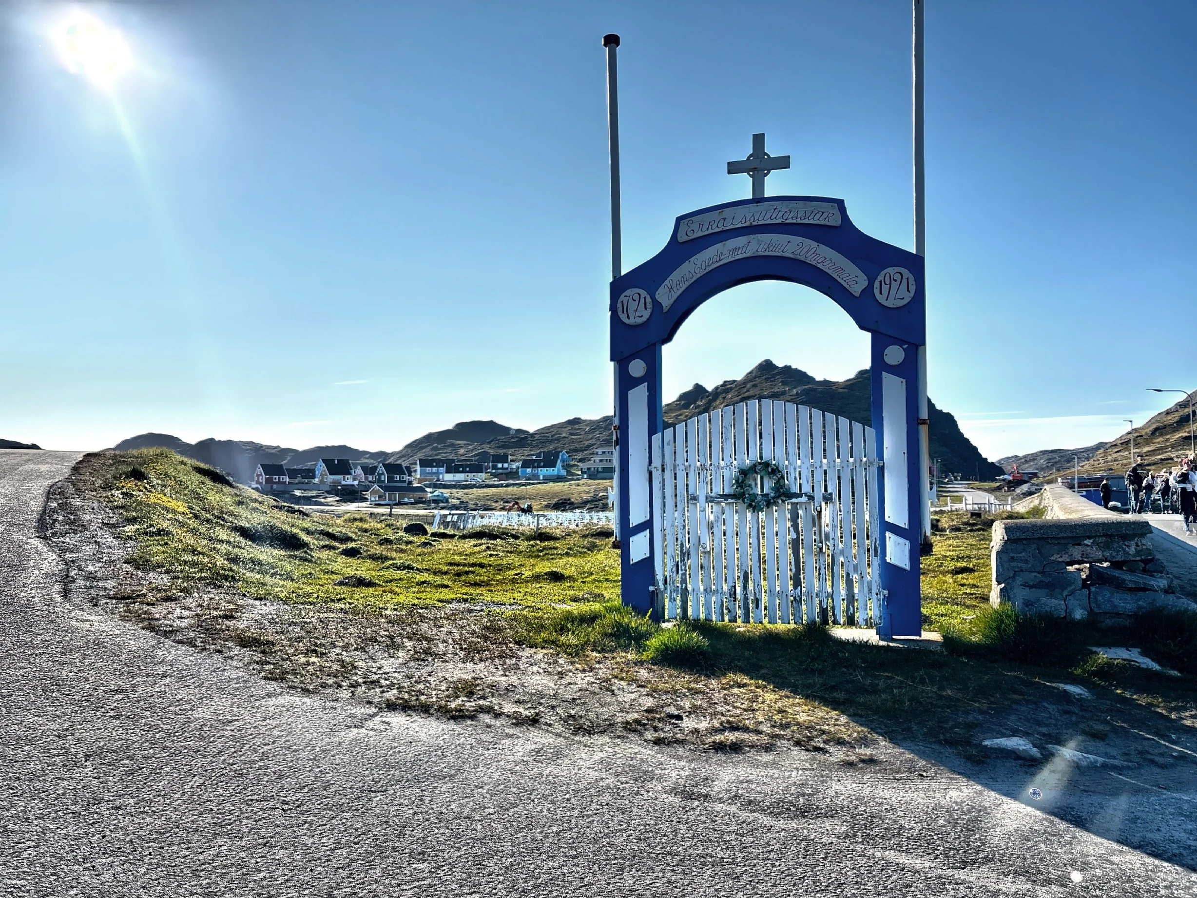  This gate marks the entrance to a peaceful cemetery, where simple white crosses rise gently from the earth, surrounded by wind-swept grass and wildflowers. 
