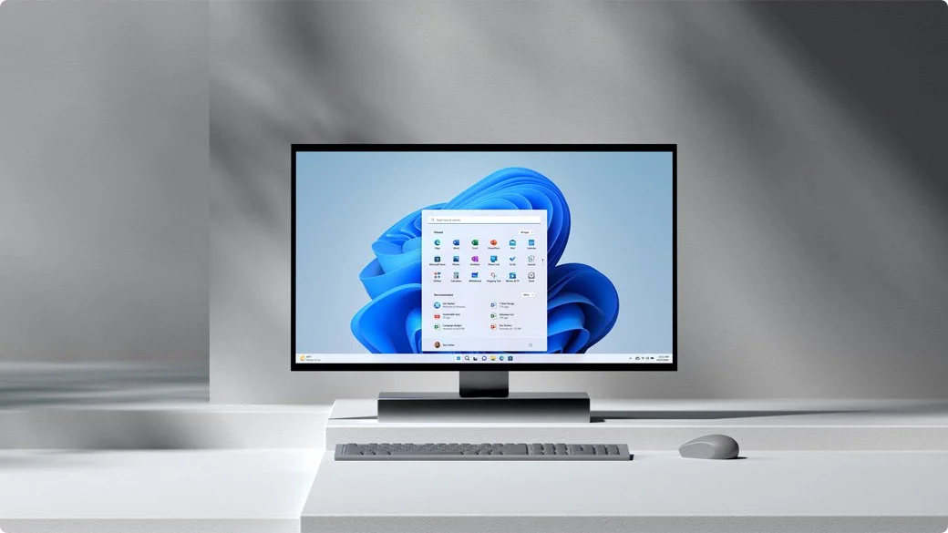 A computer monitor displaying the Windows 11 desktop with an open app menu, placed on a white desk with a keyboard and mouse.
