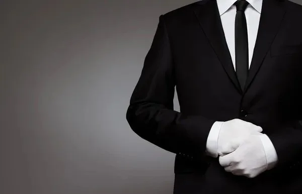 Person dressed in a black suit, white gloves, and a black tie, holding their hands together in front of them, against a plain gray background.