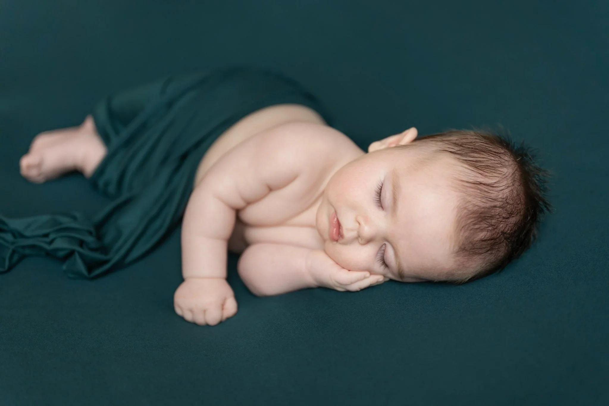 A newborn baby sleeps posed on his side on a dark teal backdrop during his Maryland newborn photography session
