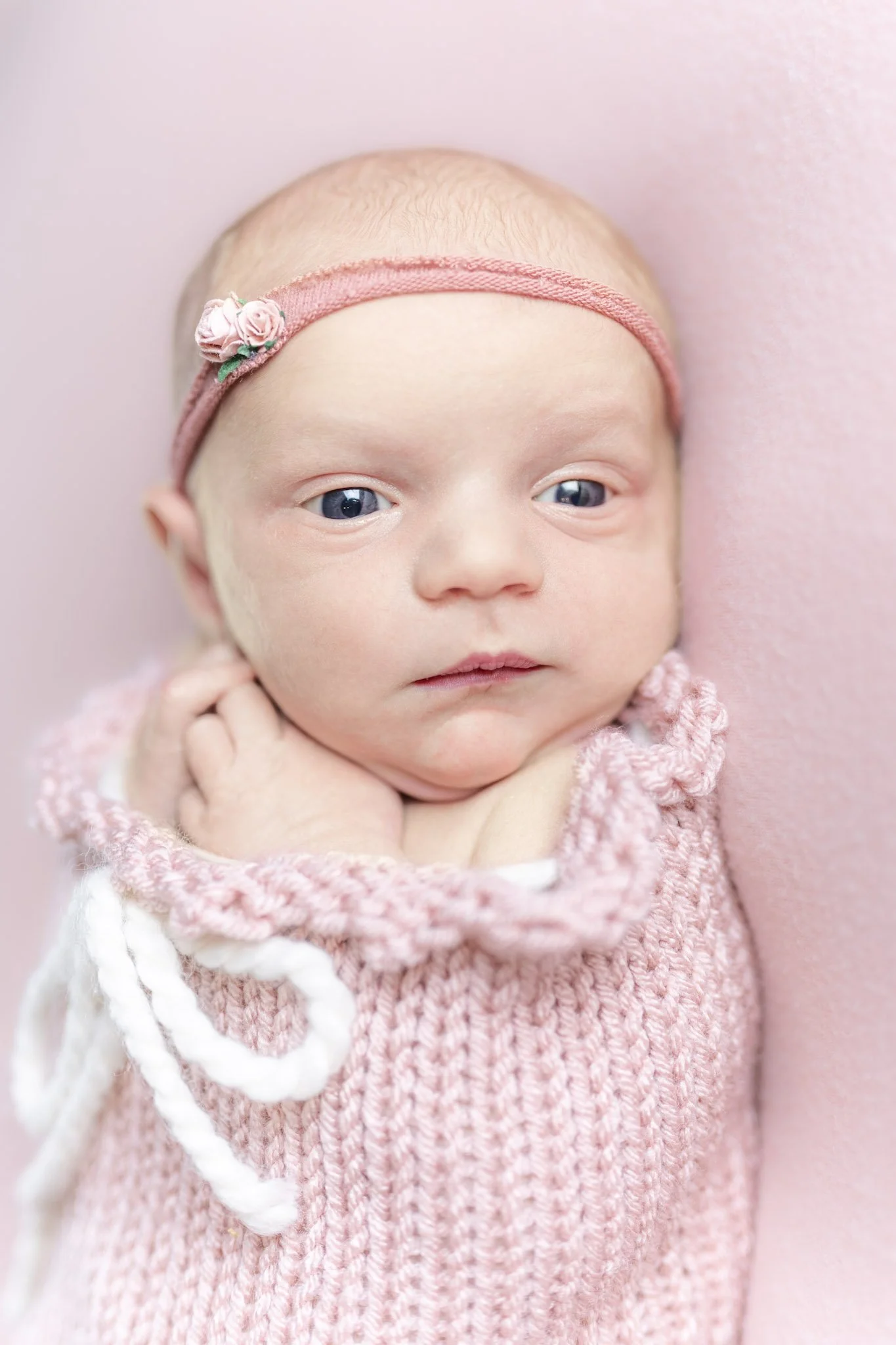 A newborn baby girl bundled up to her shoulders in a pink, thick knit sac tightened at the top by a white ribbon, wearing a pink headband with a small cluster of flowers, laying face-up on a pink backdrop during her Anne Arundel County Newborn photog