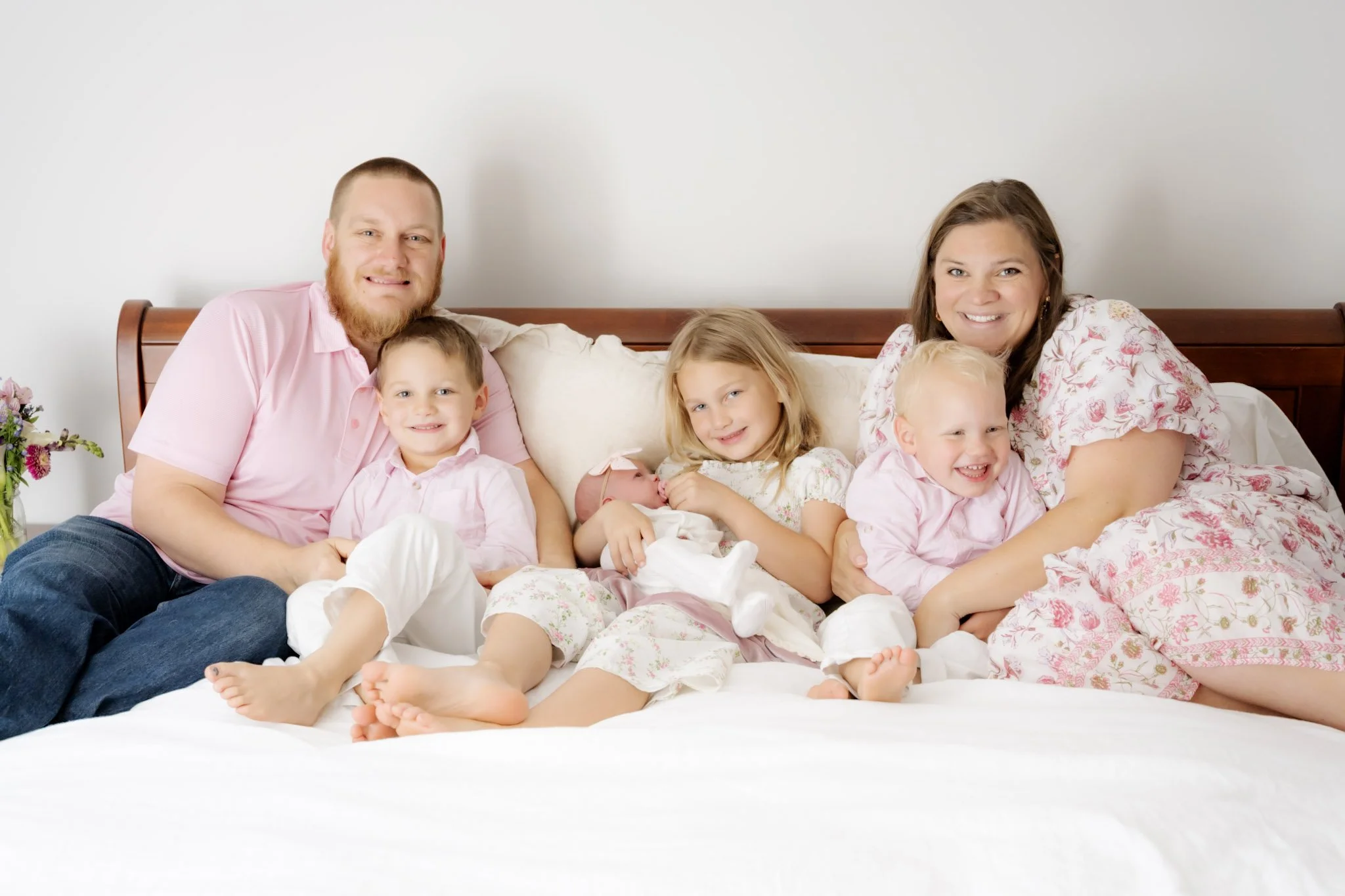 A family of 6, 2 parents and 4 kids including their newborn baby sister, sitting on a bed together posing for their Luxury Anne Arundel county Newborn Photography Session.