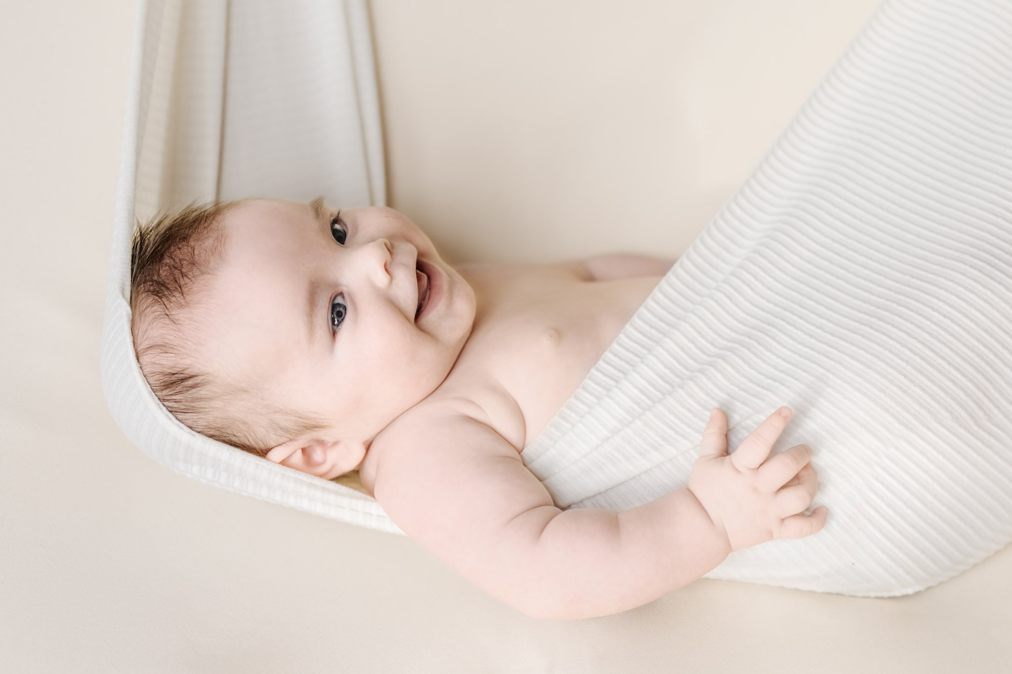A smiling newborn baby boy lies in a makeshift "hammock" fashioned from a cream-colored baby wrap hanging in front of a beige backdrop during his Anne Arundel County newborn photography session.