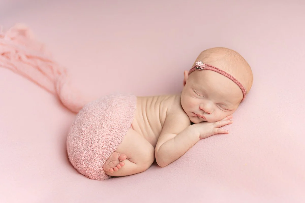A newborn baby sleeping on a pink backdrop, wearing a pink lace gauze wrap and pink flower headband during her Crownsville Newborn Photography session