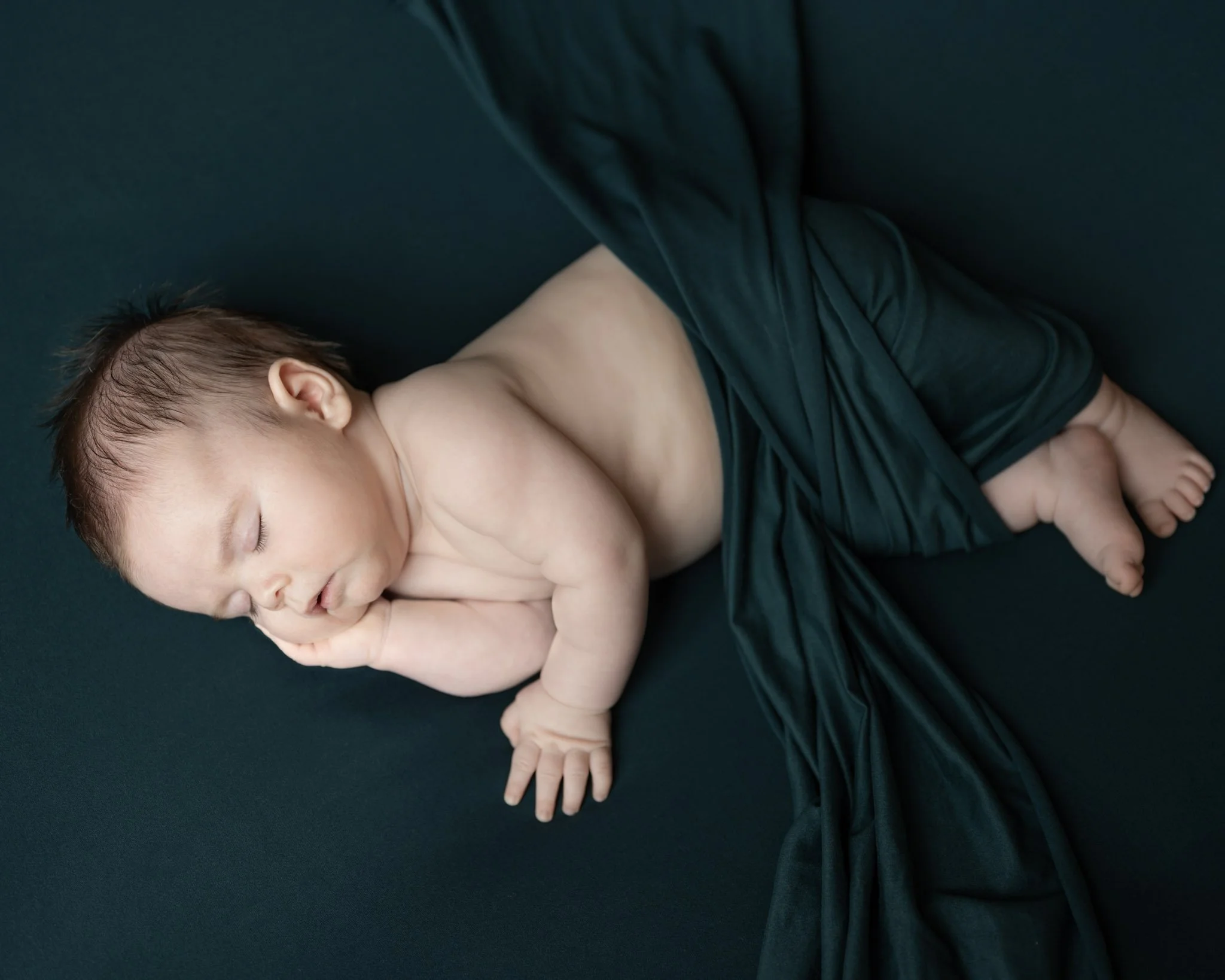 A newborn baby boy sleeping on his side with one hand under his cheek for his Anne Arundel County newborn photography session is wrapped in a dark turquoise wrap and is laying on a matching backdrop. 