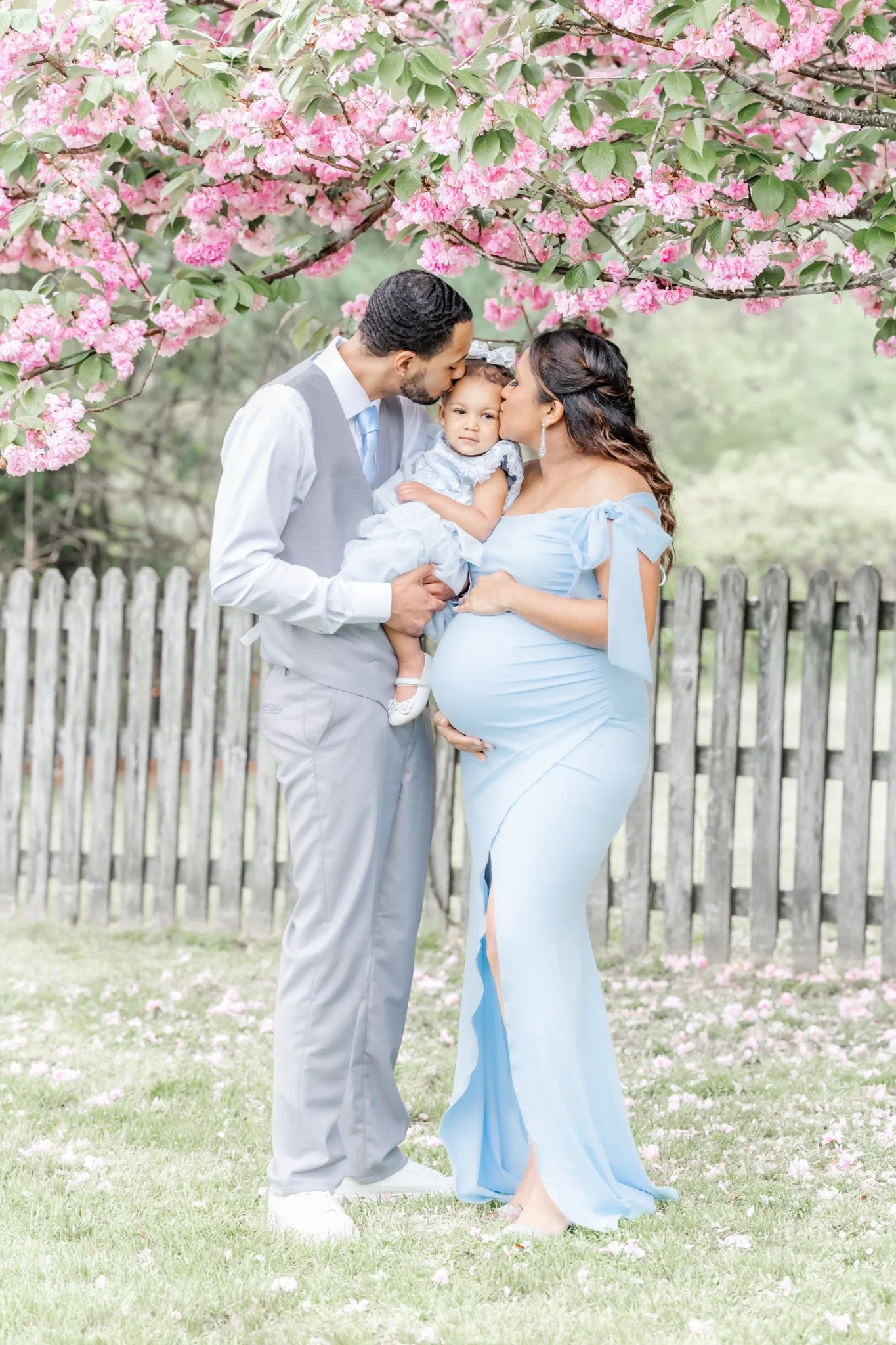 A pregnant mom and dad stand holding their toddler daughter between them each giving her a kiss on the head during their maternity photography session in Severna Park, Maryland
