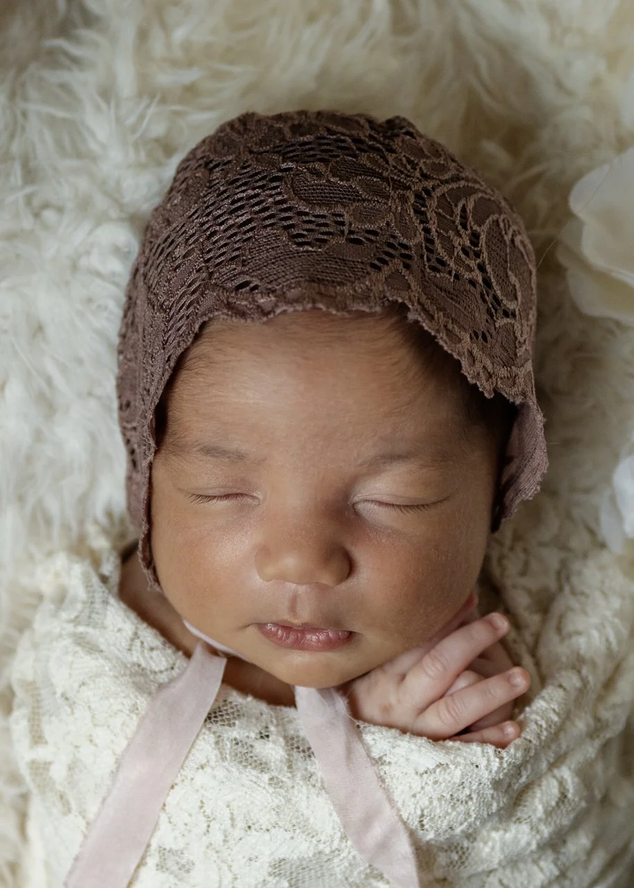 A newborn baby wrapped in white lace, wearing a dark dusty red-pink lace bonnet sleeps on a white flokati during her Maryland Newborn photography session.
