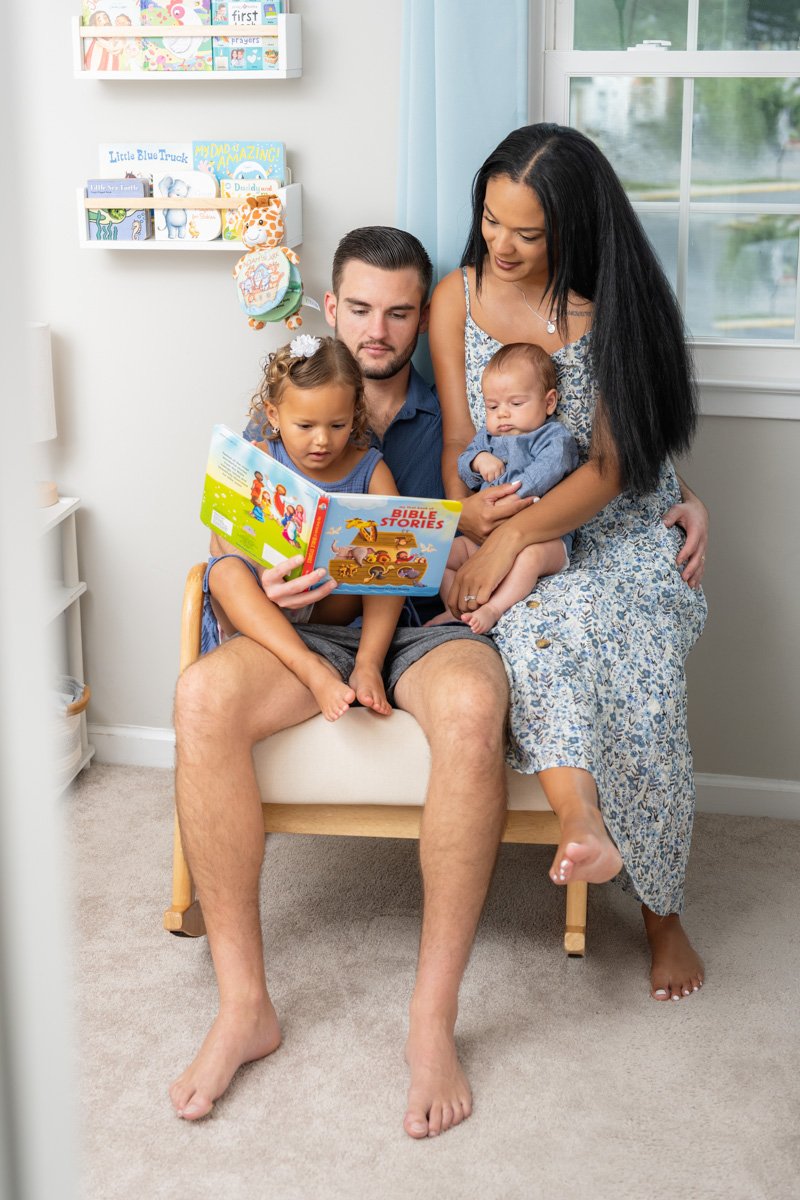 A newborn baby sits with his parents and sister in it's nursery while they read a book during his Maryland newborn photography session.