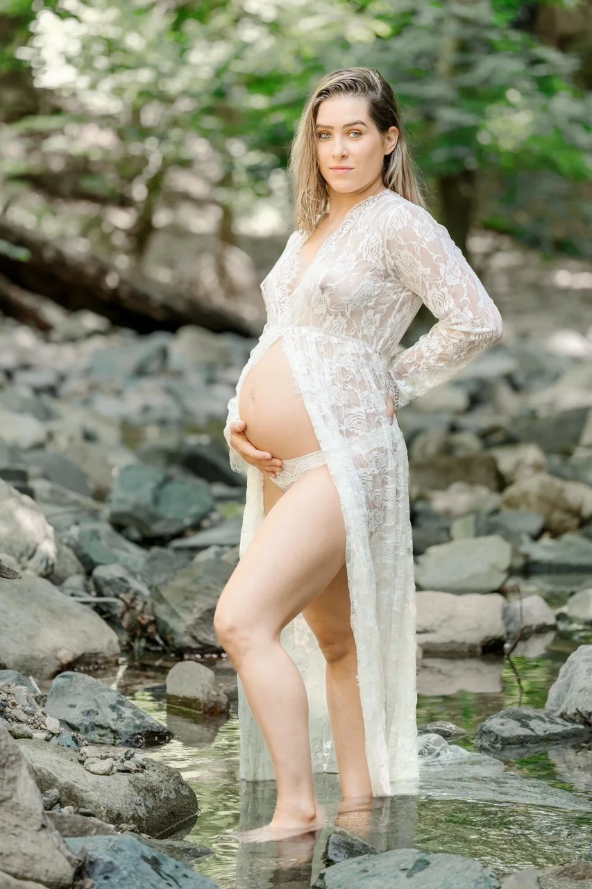 A pregnant woman in a sheer lace robe standing in a shallow creek surrounded by rocks and greenery.