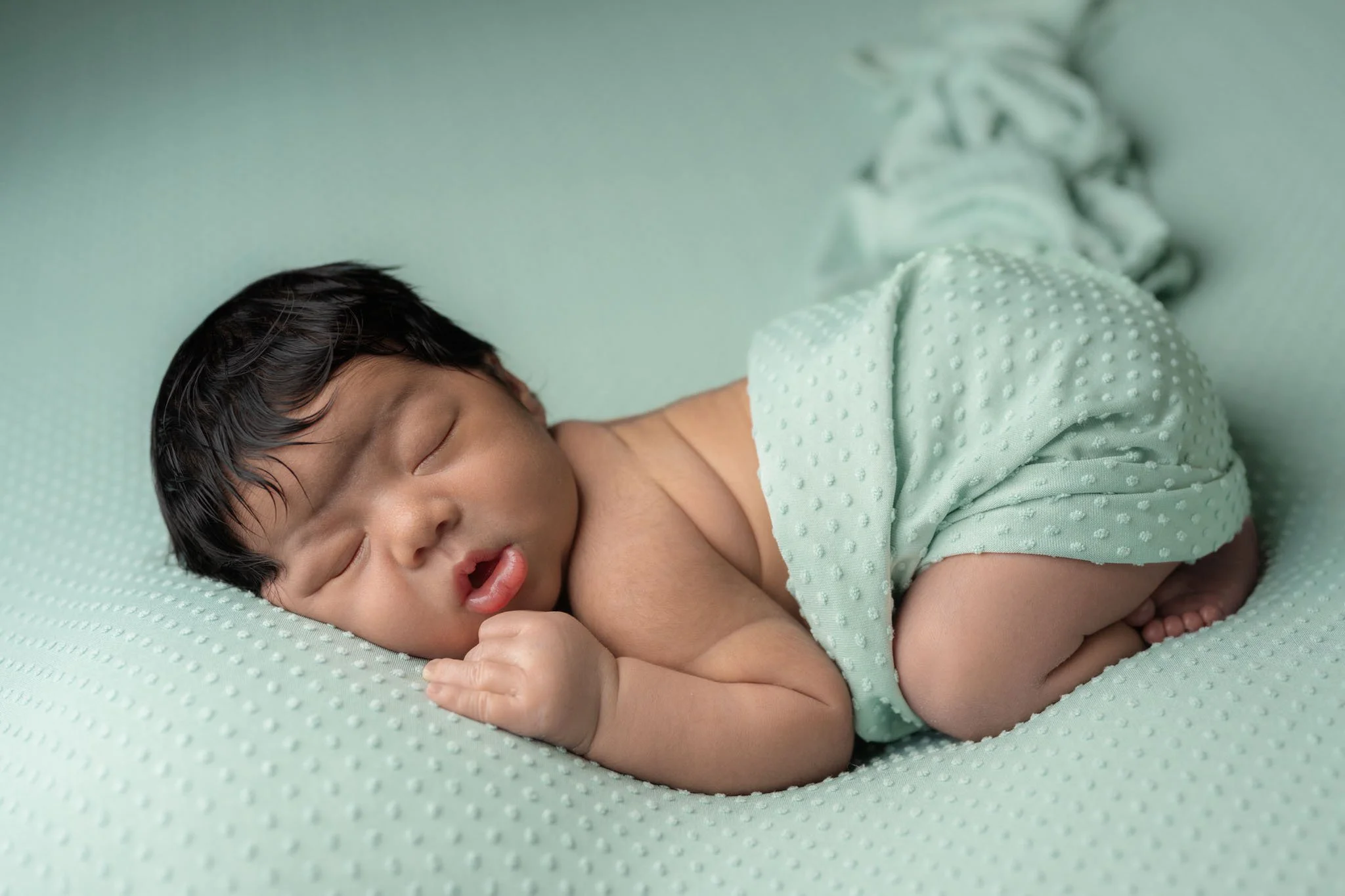 A newborn baby boy wrapped in a light turquoise wrap, with jet black hair, sleeps curled up on his belly with his legs tucked under him on a light turquoise backdrop during his newborn photography session in Bowie, MD.