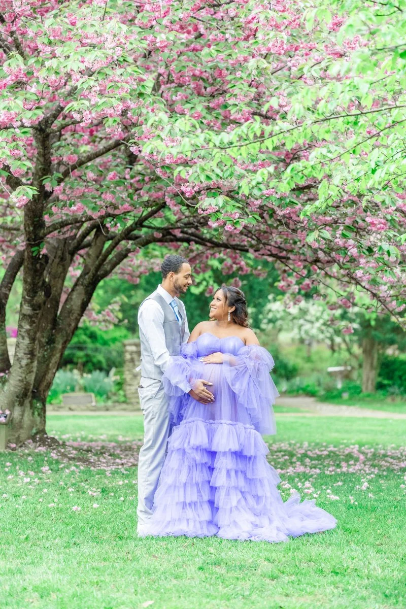 An expectant couple standing under a blooming tree, both touching her pregnant belly. The mother is wearing a flowing purple gown. The father is wearing a grey suit during their Crownsville Maternity photography session.