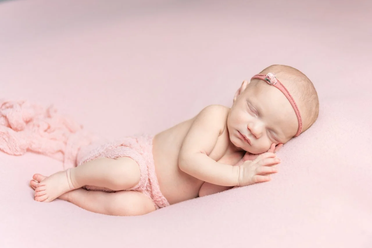 A sleeping newborn baby with a pink headband and pink wrap, lying on a soft pink surface during her Anne Arundel County Newborn Photography session