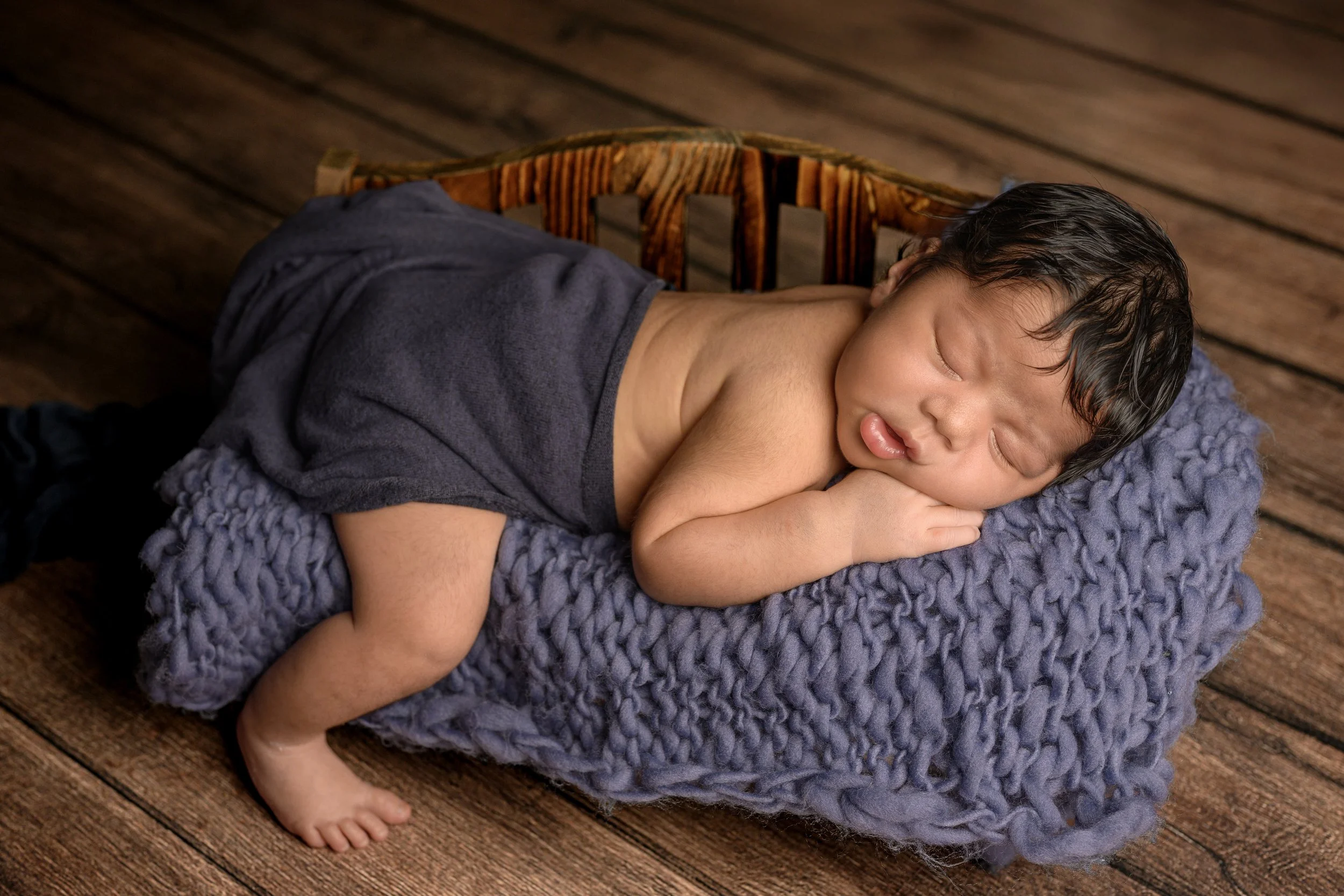 A newborn baby boy sleeping on his tummy during his Anne Arundel County newborn photography session, hands folded, one on top of the other under his cheek, he lays atop a navy blue chunky knit blanket with a dark navy-blue wrap covering his diaper.