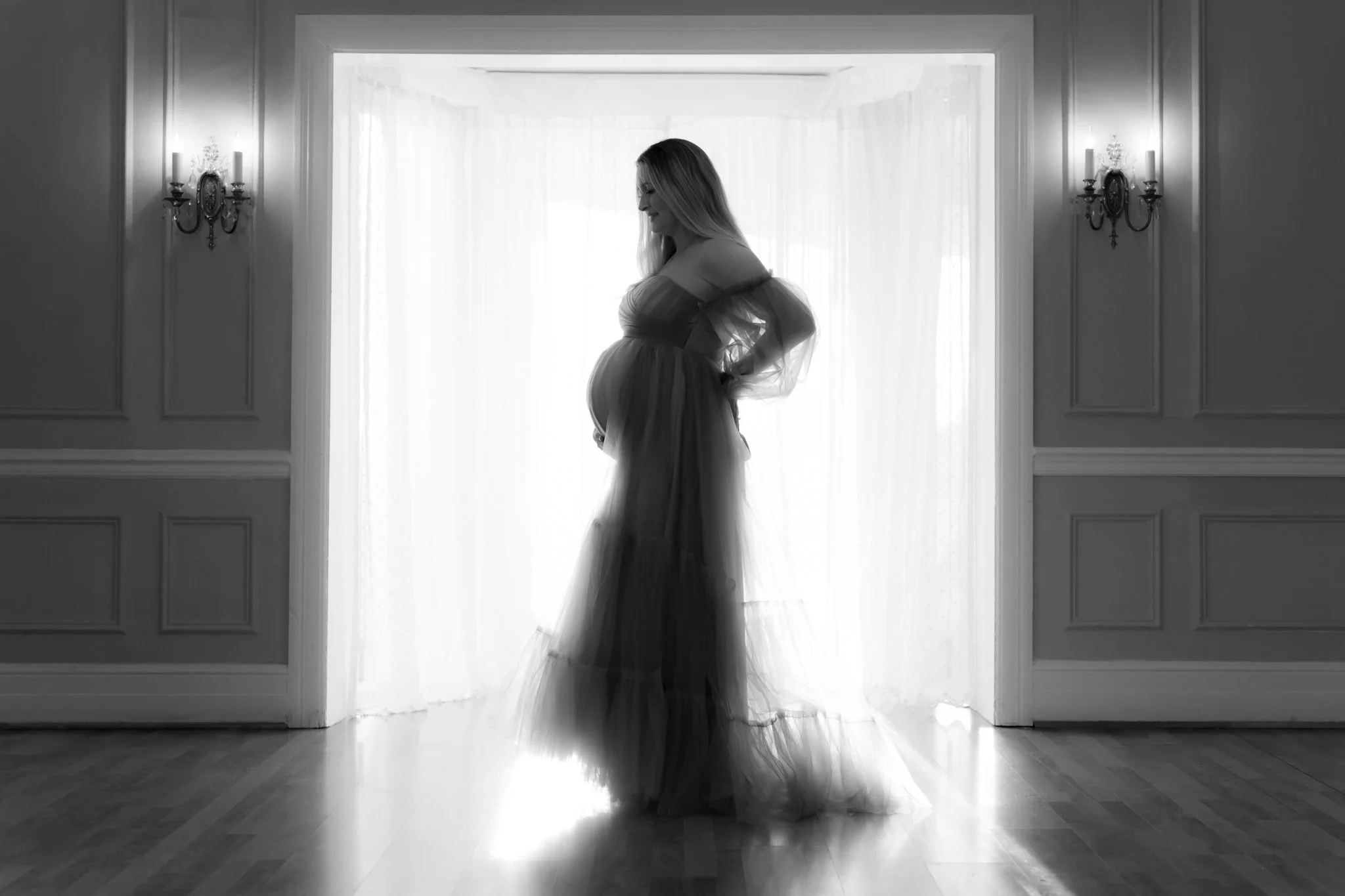 A black and white photo of a pregnant woman in a flowing tulle gown standing in front of a bright window causing her to be in silhouette during her Annapolis Maternity photography session