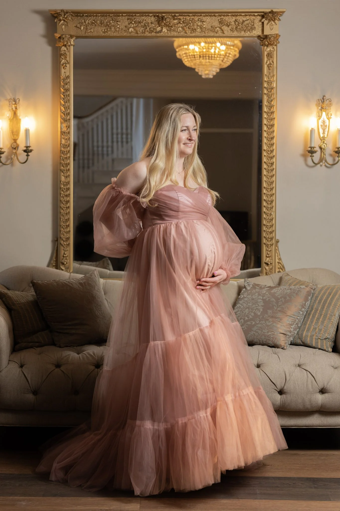A pregnant mother in a mauve, tulle, flowing gown stands in a dimly lit room with her hand on her belly awaiting her Crownsville Newborn photography session.