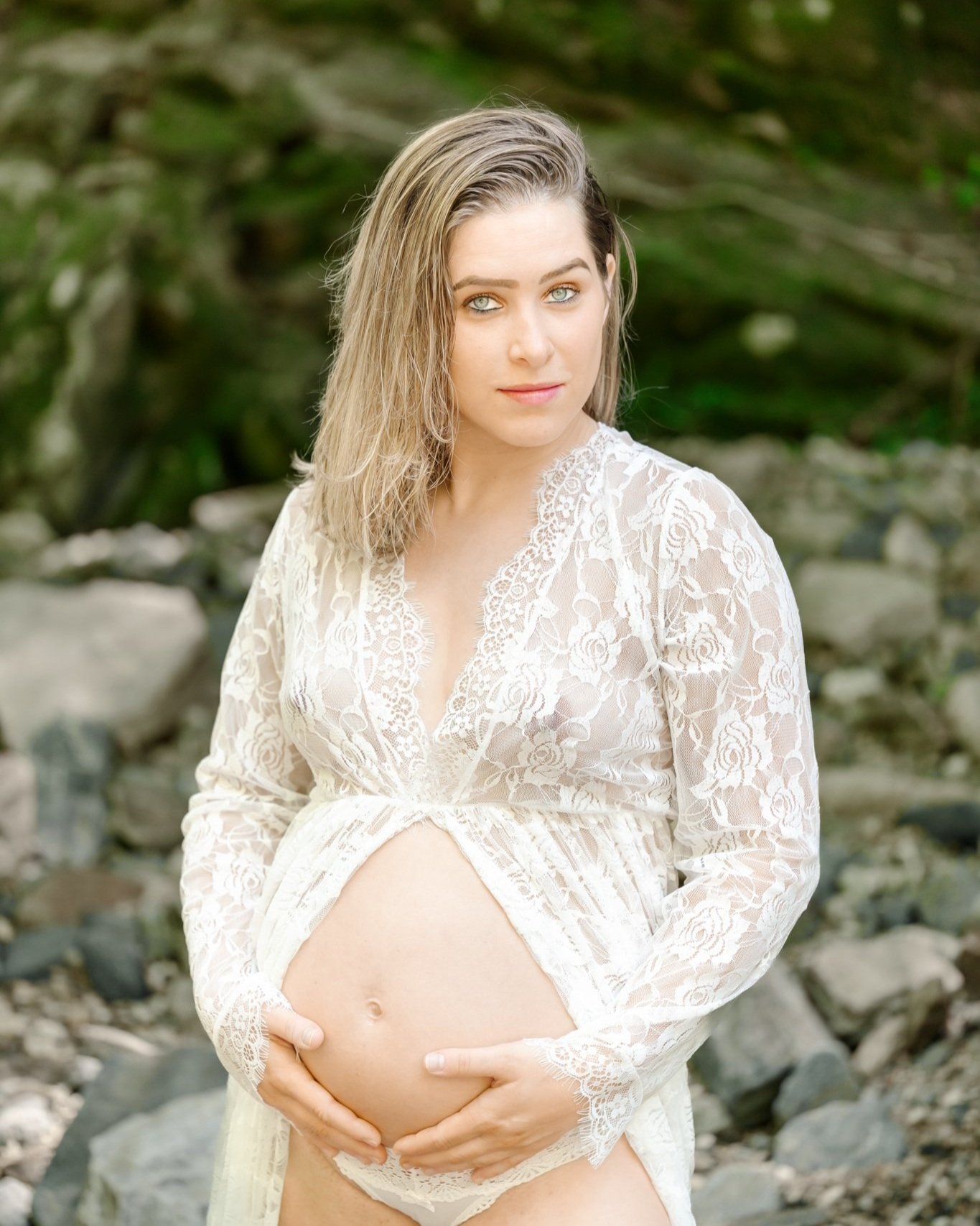 A pregnant woman wearing a shear lace duster, open in the front to expose her belly, holds her hands on her belly and gives a piercing look at the camera during her boudoir, maternity photography session in Maryland.