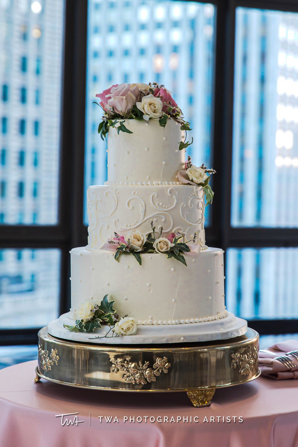 Three-tier white wedding cake decorated with flowers on a silver stand, set on a pink tablecloth with a cityscape background inside a building.