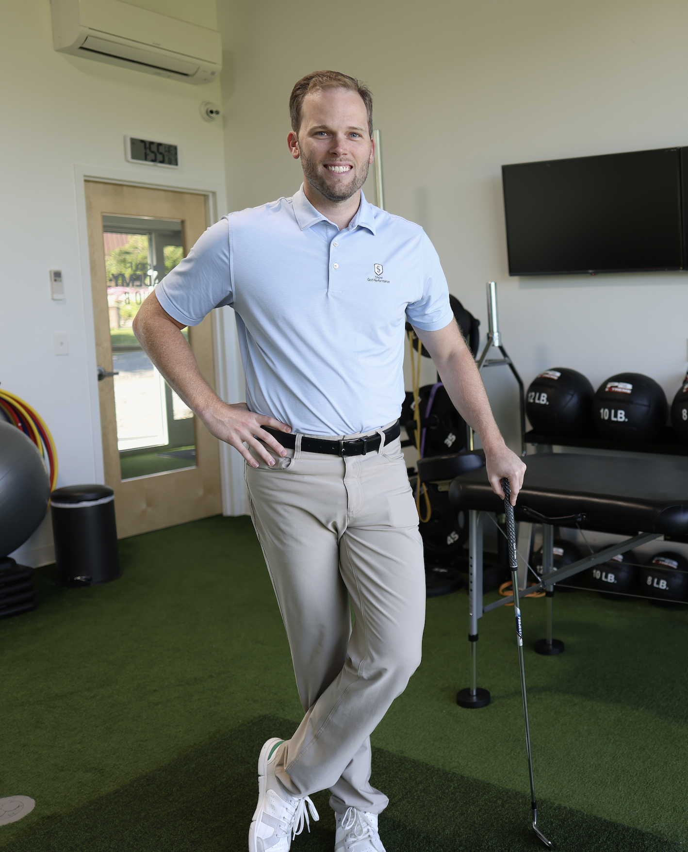 A man in golf attire standing in a gym, holding a golf club, with fitness equipment in the background.