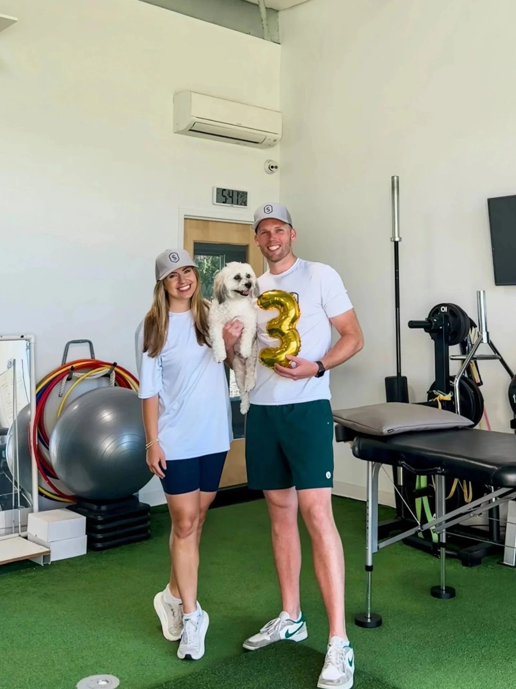 A smiling couple holding a dog and a gold balloon shaped like the number 3, standing inside a gym or physical therapy clinic.
