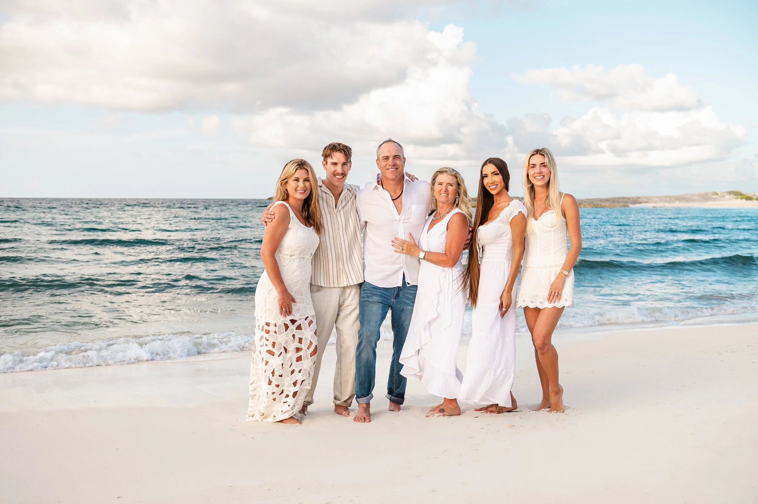 Family of five standing on a beach, smiling, with the ocean and cloudy sky in the background.