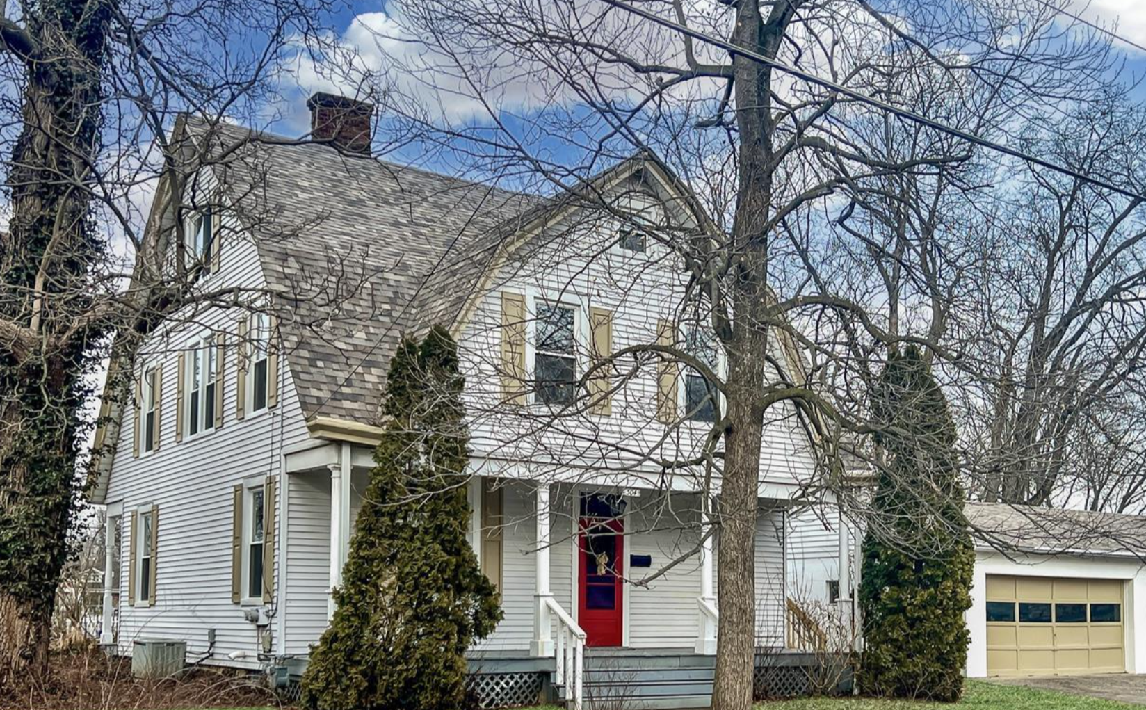 White two-story house with a red front door, surrounded by leafless trees, and an attached garage with a beige door, under a partly cloudy sky.