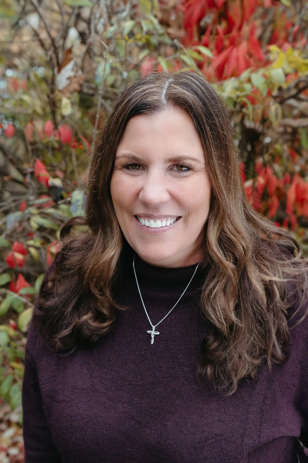 A woman smiling outdoors with autumn foliage in the background.