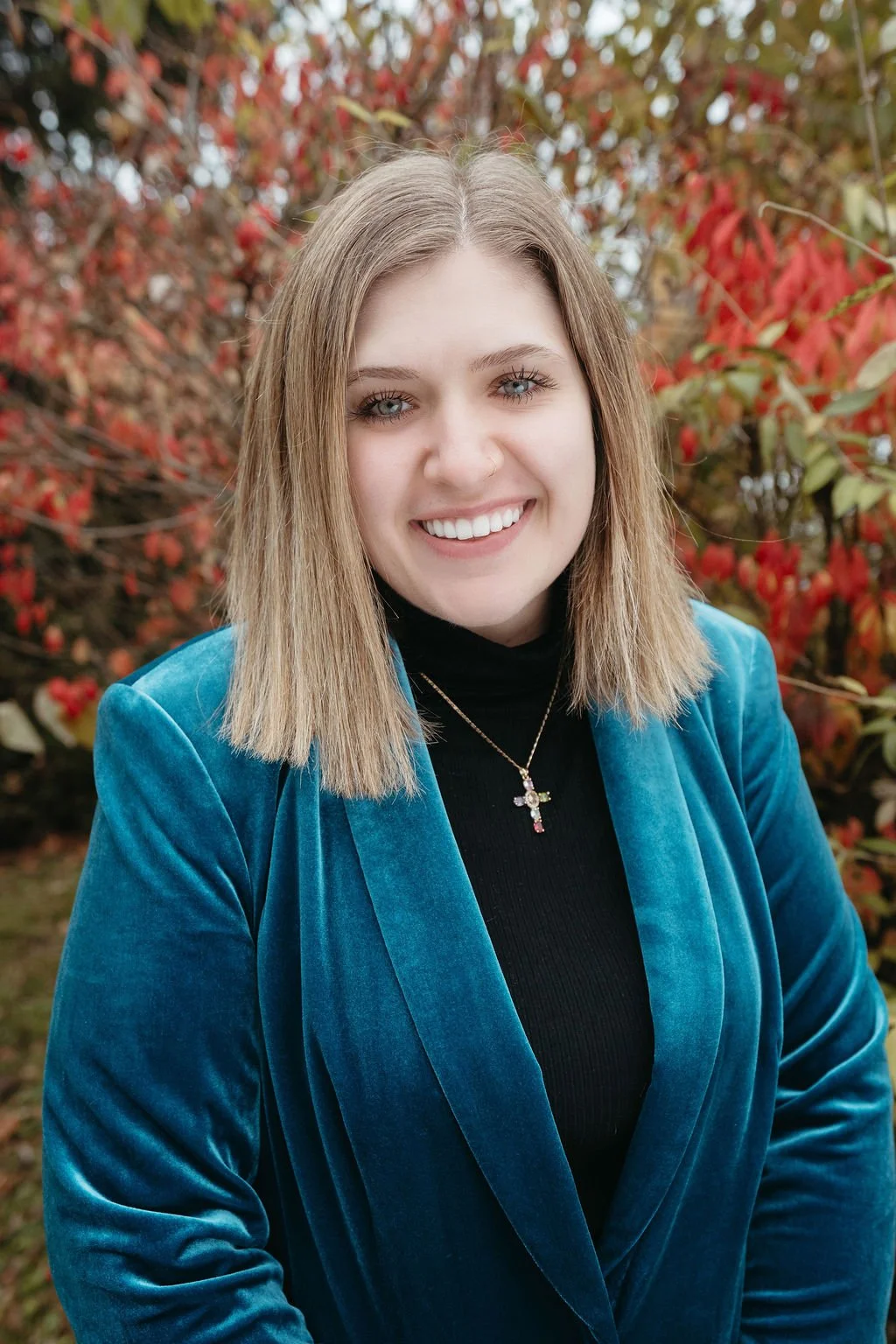 A young woman with blonde hair and blue eyes smiling outdoors with a background of red and green autumn leaves. She is wearing a black turtleneck, a blue velvet blazer, and a gold necklace with a cross pendant.