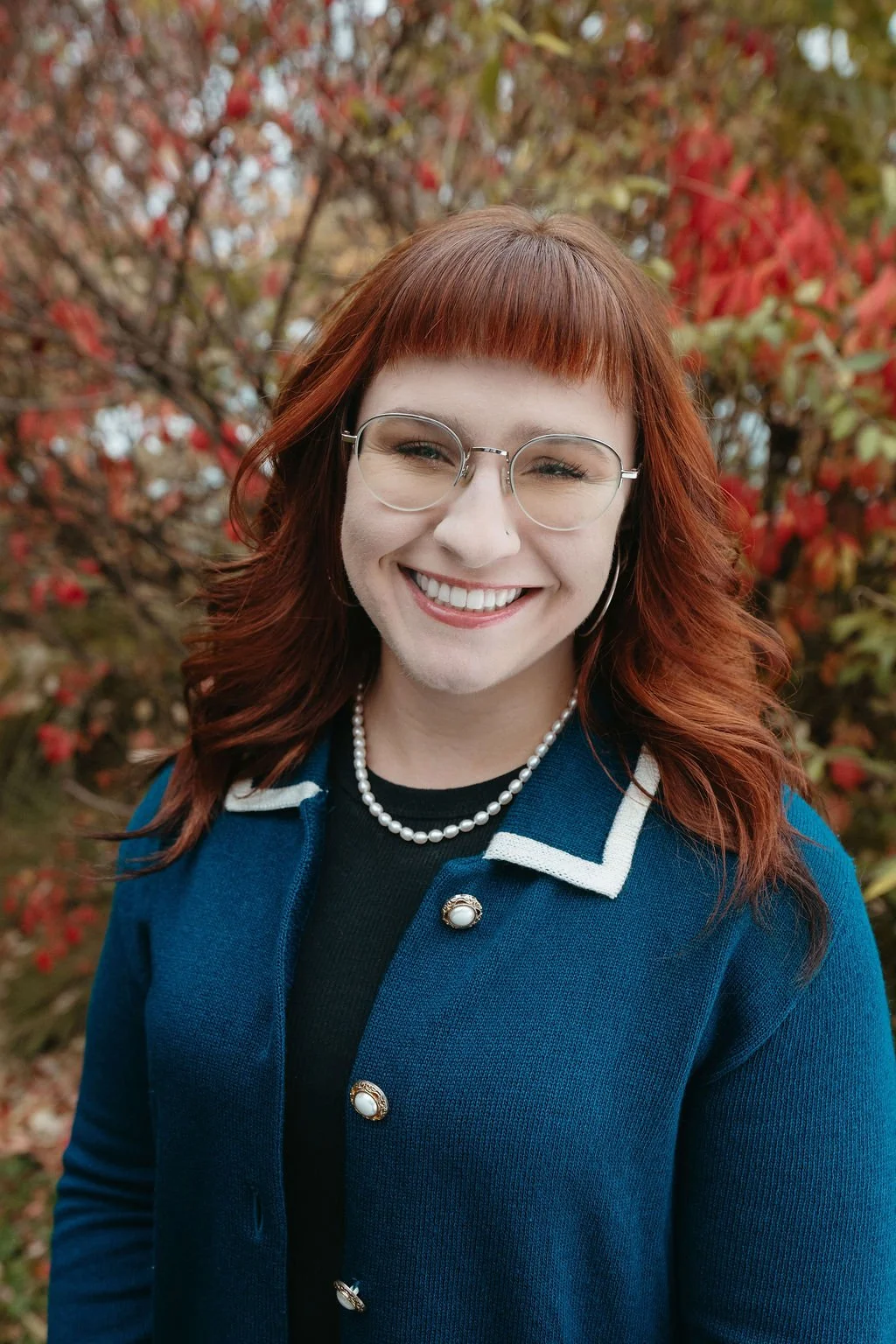 A young woman with red hair, glasses, and a pearl necklace, smiling outdoors in front of autumn foliage.