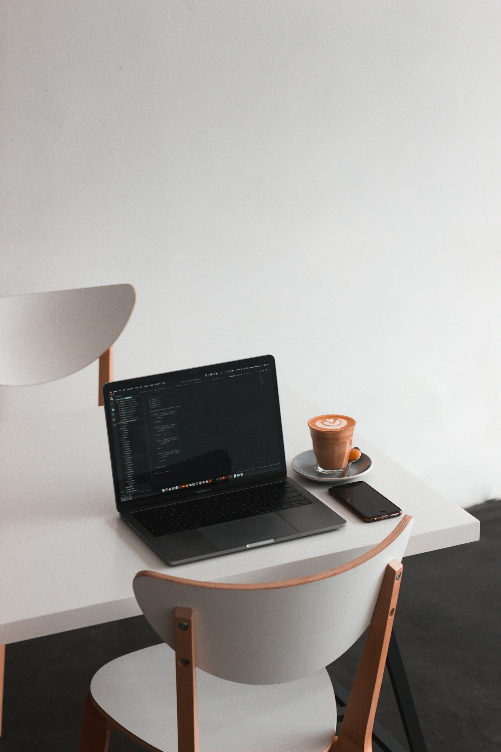 A minimalistic workspace featuring a white desk with a MacBook displaying lines of code, a smartphone, and a cup of coffee with latte art on a saucer, set against a plain white wall.