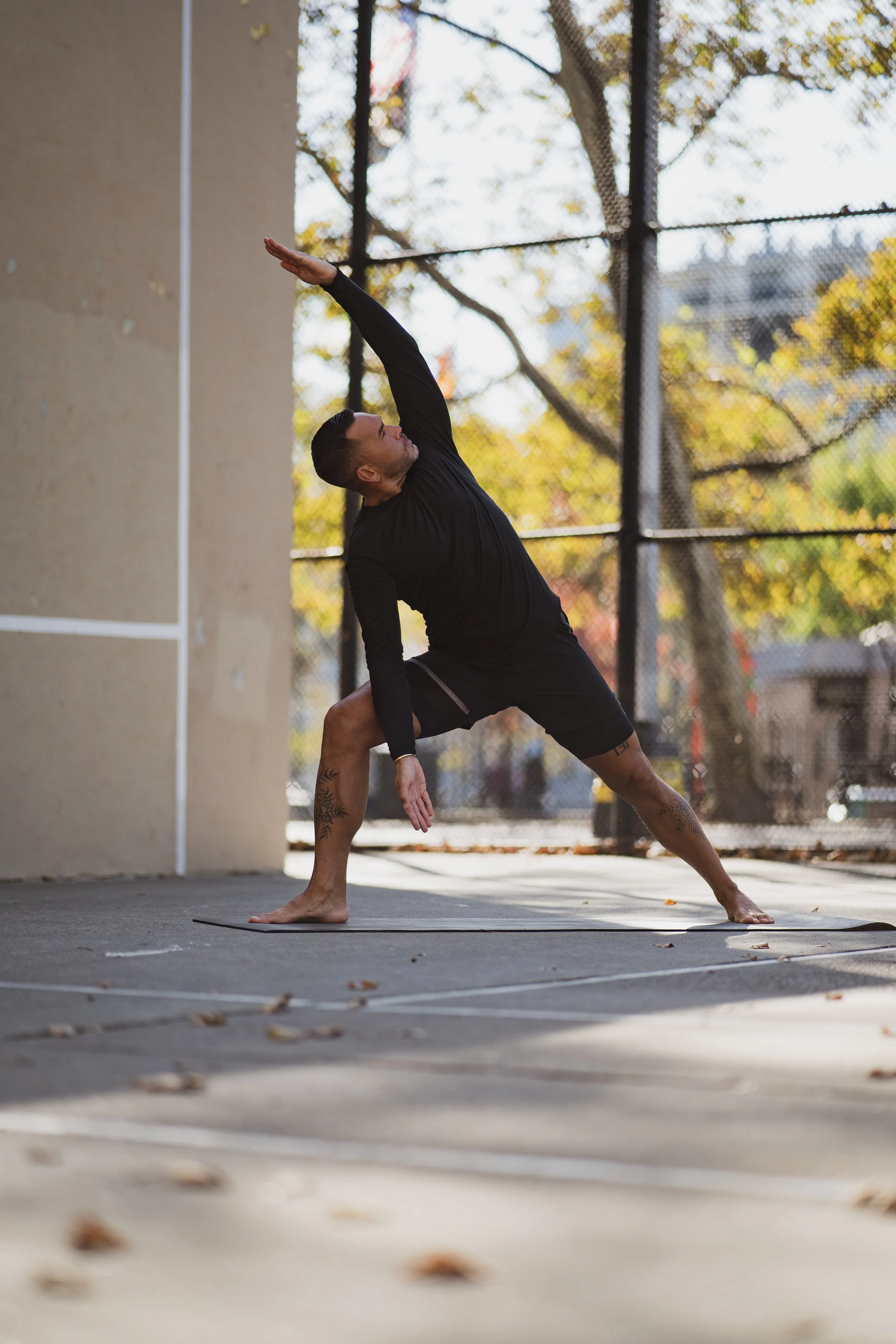 A man practicing yoga outdoors on a concrete court surrounded by a chain-link fence, with autumn trees in the background, wearing black athletic clothing, in a wide-legged pose.