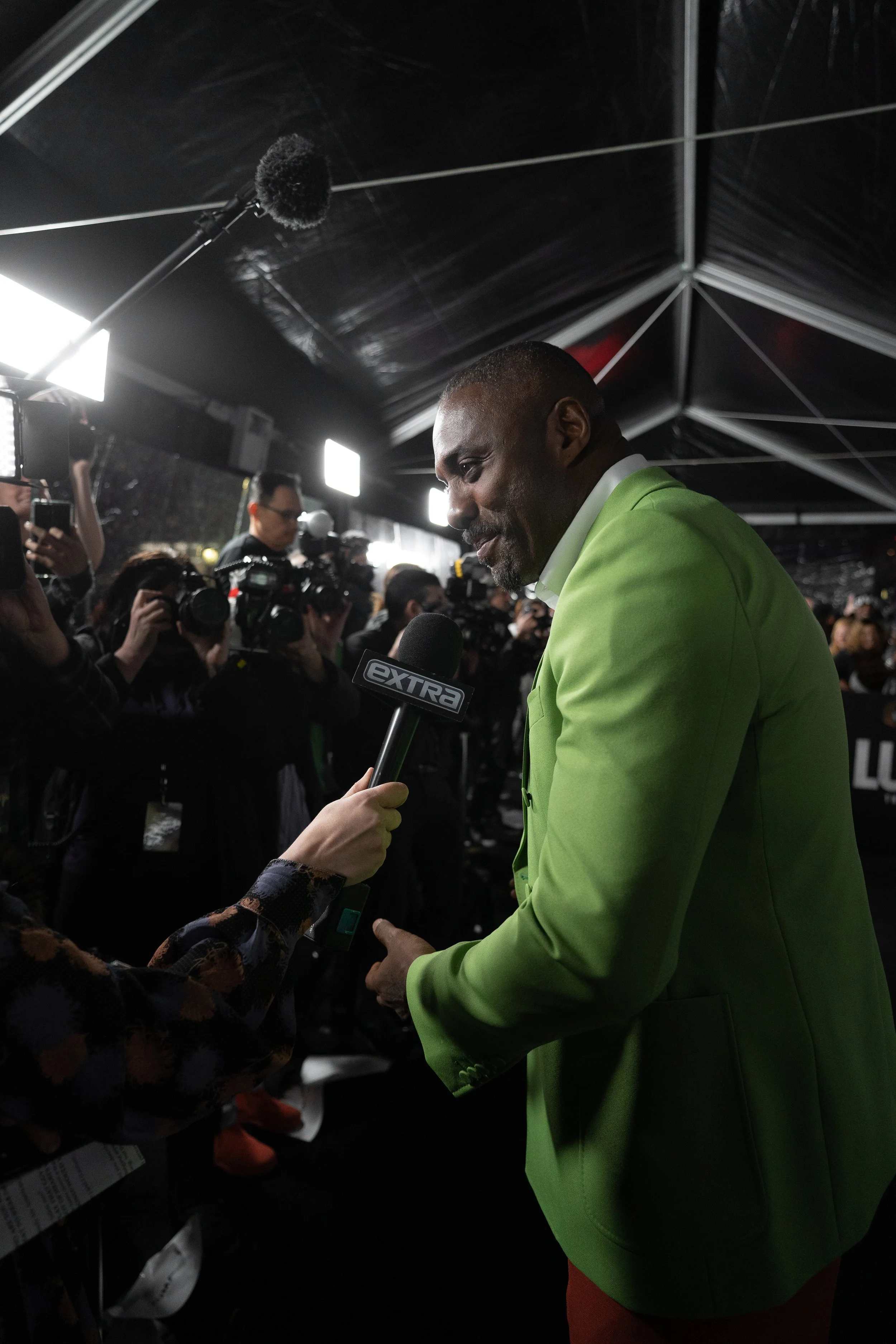 Man in bright green blazer giving an interview to a microphone held by a person with a patterned sleeve, surrounded by photographers and cameras at a media event.