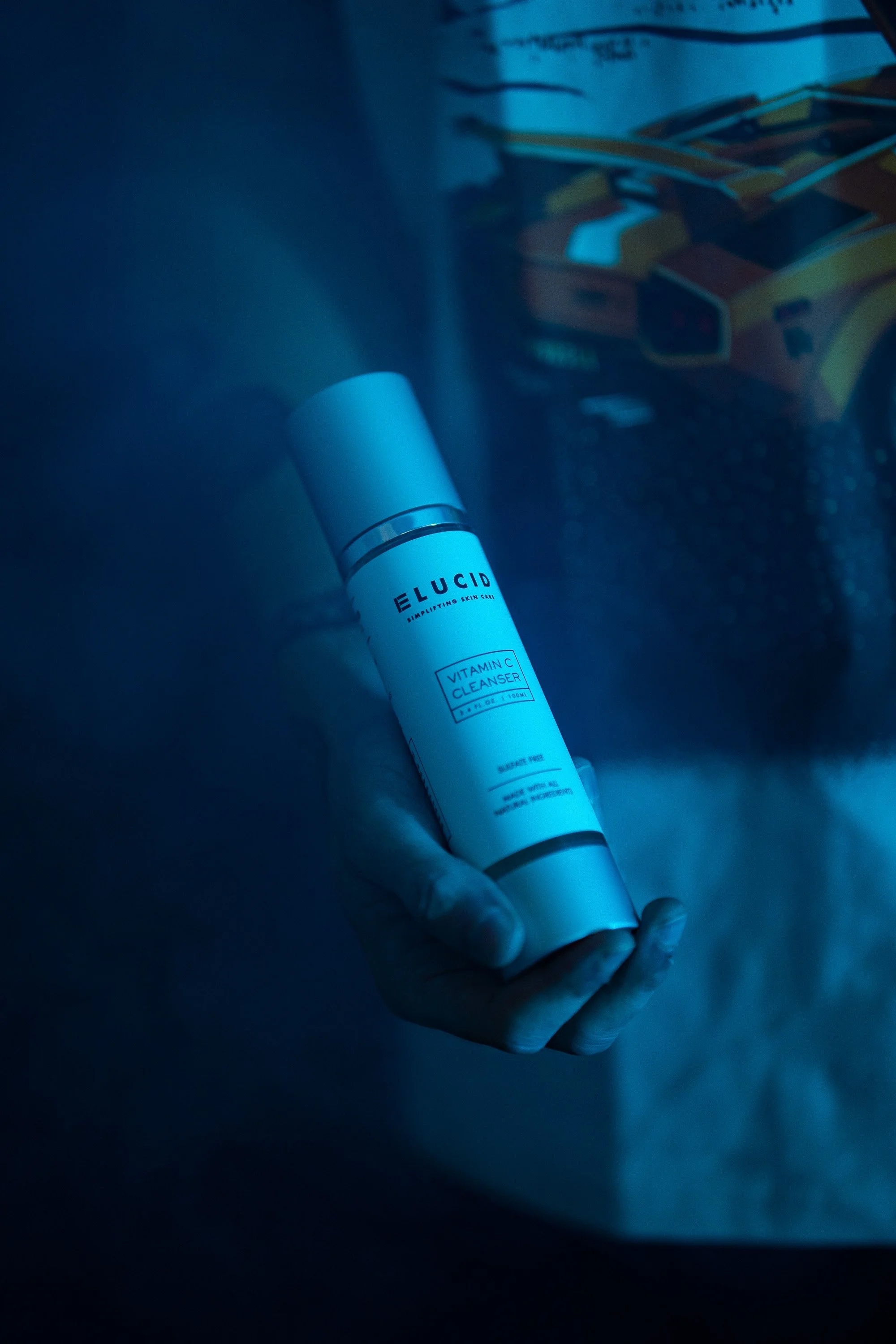 A hand holding a white bottle of Elucid Vitamin C Cleanser in a dimly lit room with a toy car in the background.