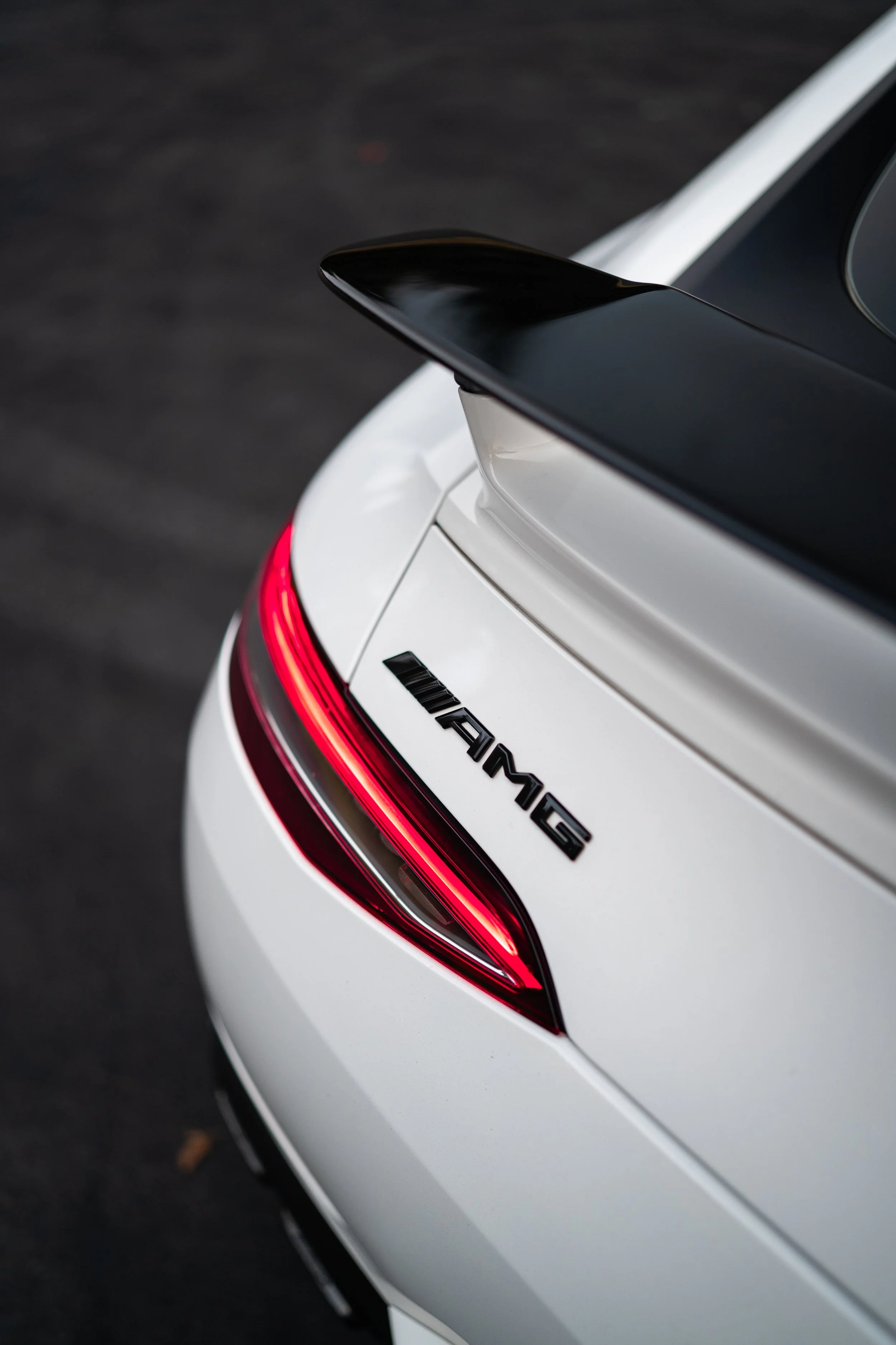 Close-up of the rear end of a white Mercedes-AMG sports car, featuring a black spoiler, red tail lights, and AMG badge.