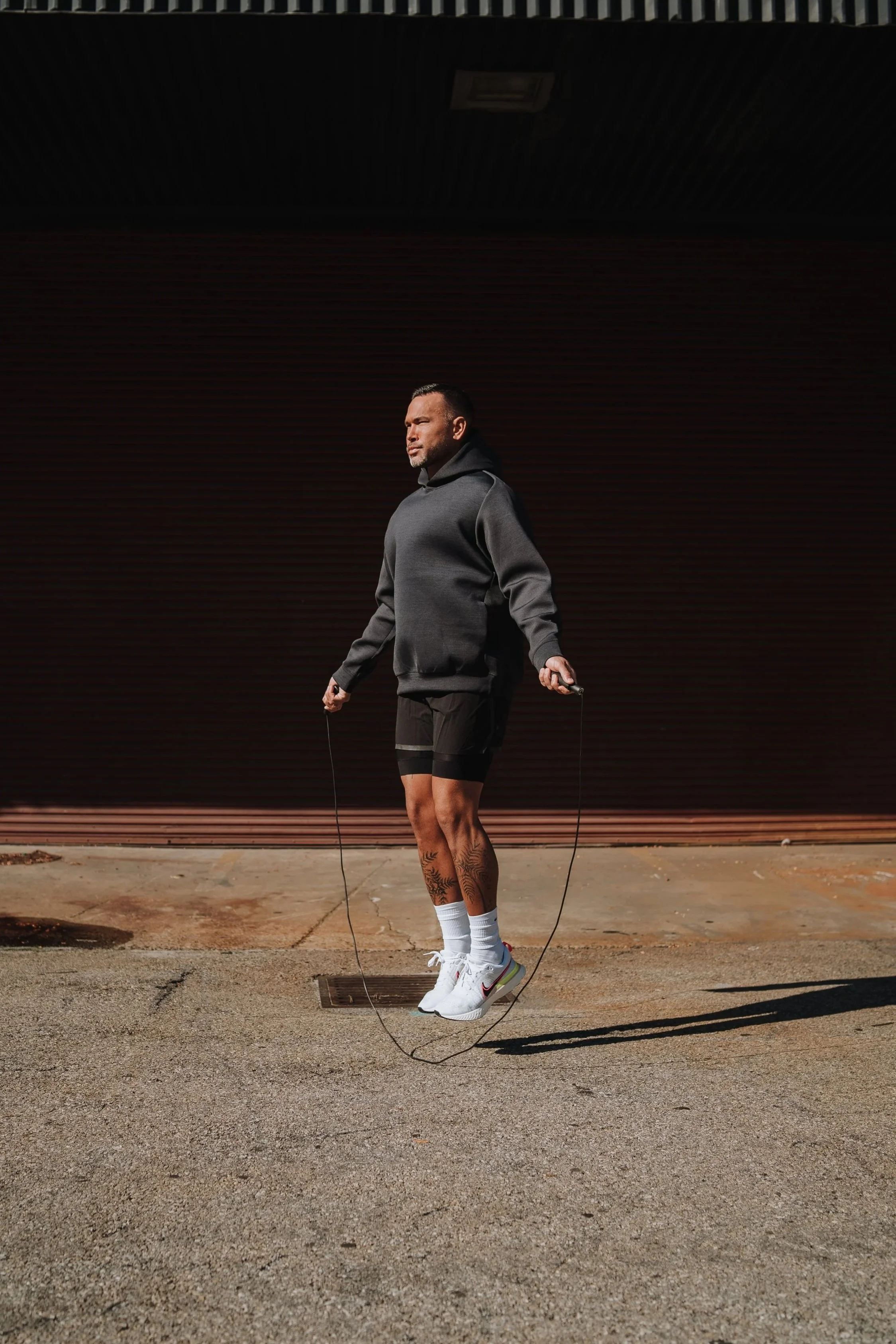 A man in athletic clothing, including shorts, sneakers, and a hoodie, is jumping rope outdoors on a concrete surface near a closed, brown industrial garage door, with shadows cast on the ground.