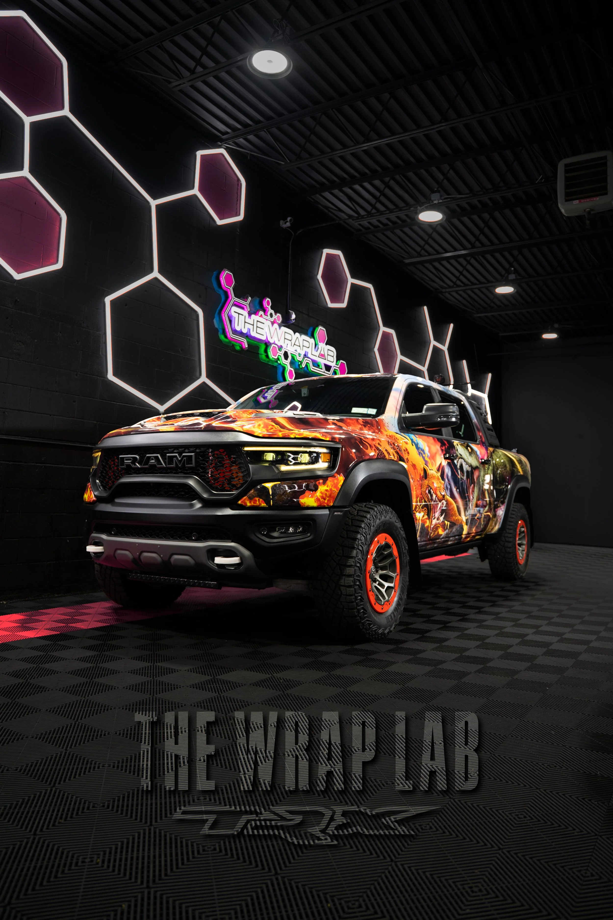 A customized RAM pickup truck with a fiery graphic wrap on its body, parked indoors on a checkered floor. The background features a black wall with hexagonal light fixtures and neon signs that read 'THE WRAP LAB' with a colorful logo.