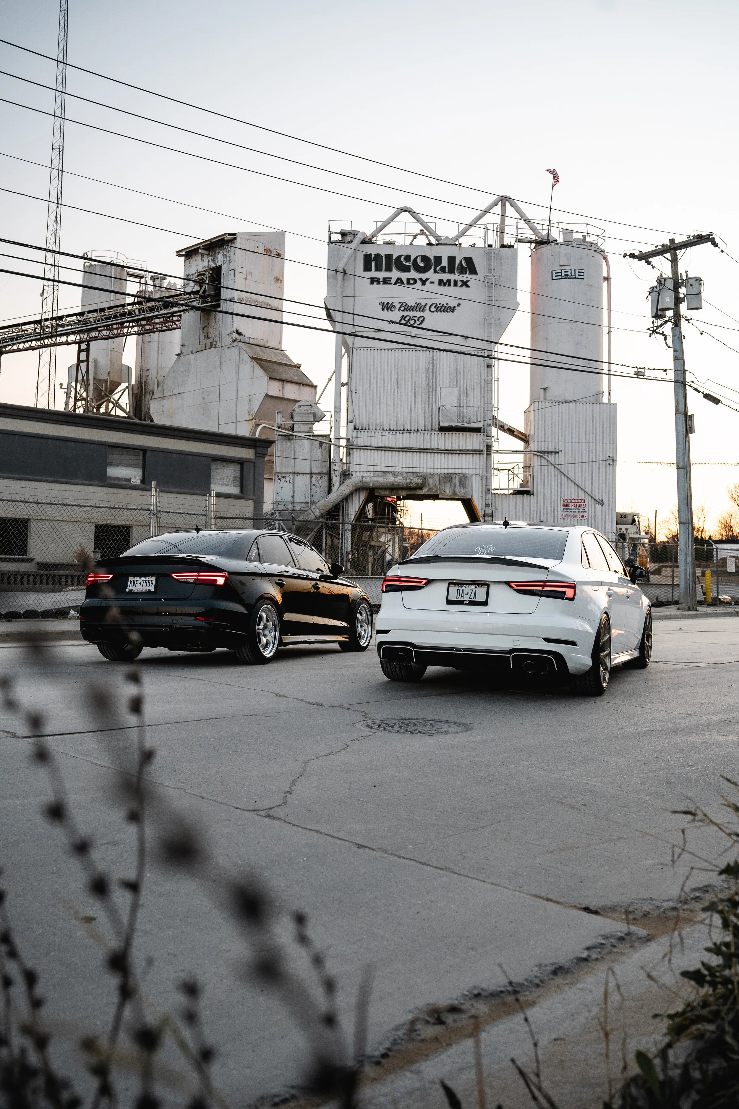 Two modern cars parked in an industrial area with factory silos and power lines in the background during sunset.