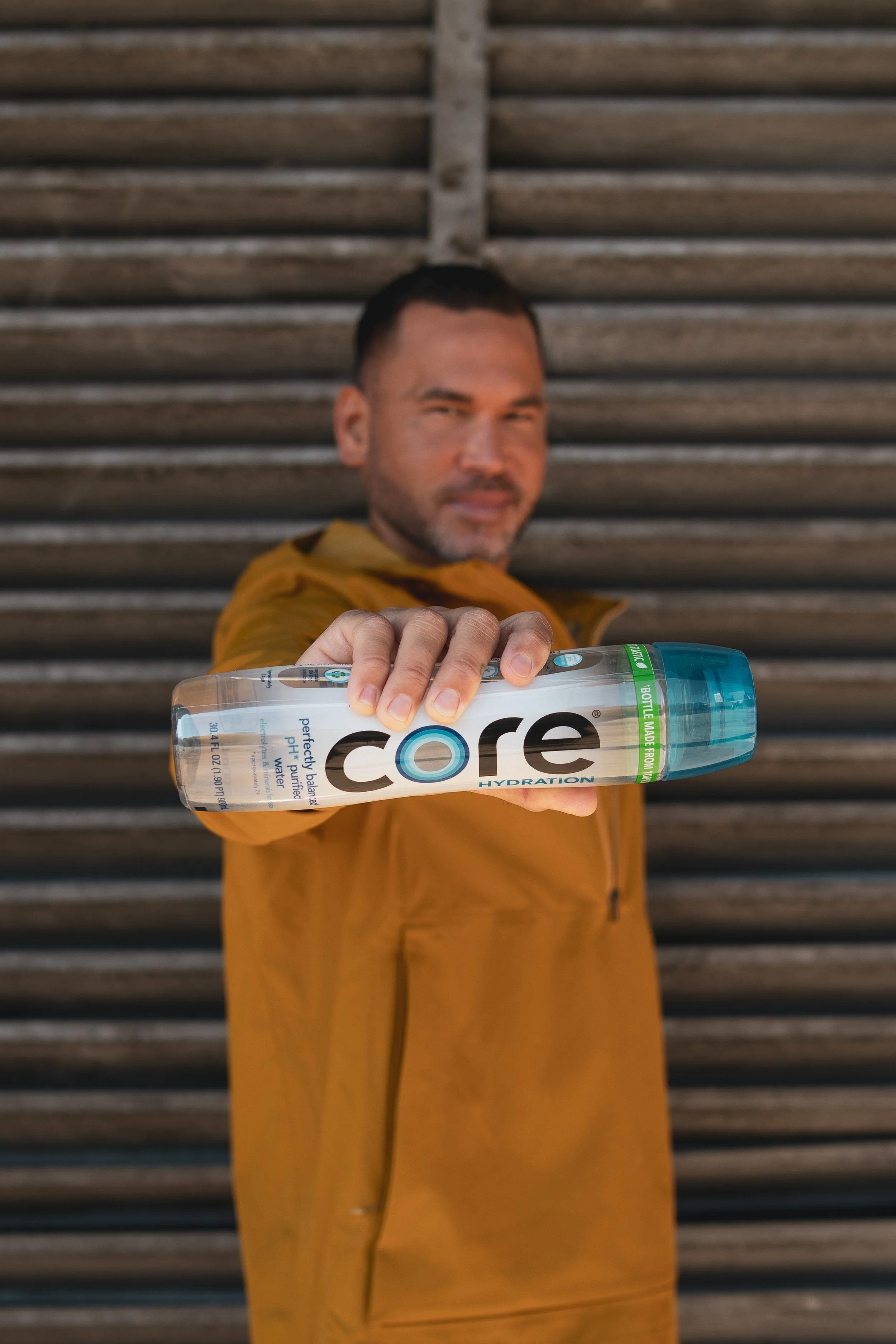 Man in yellow jacket holding a clear water bottle labeled 'core hydration' directed toward the camera with a wooden background.