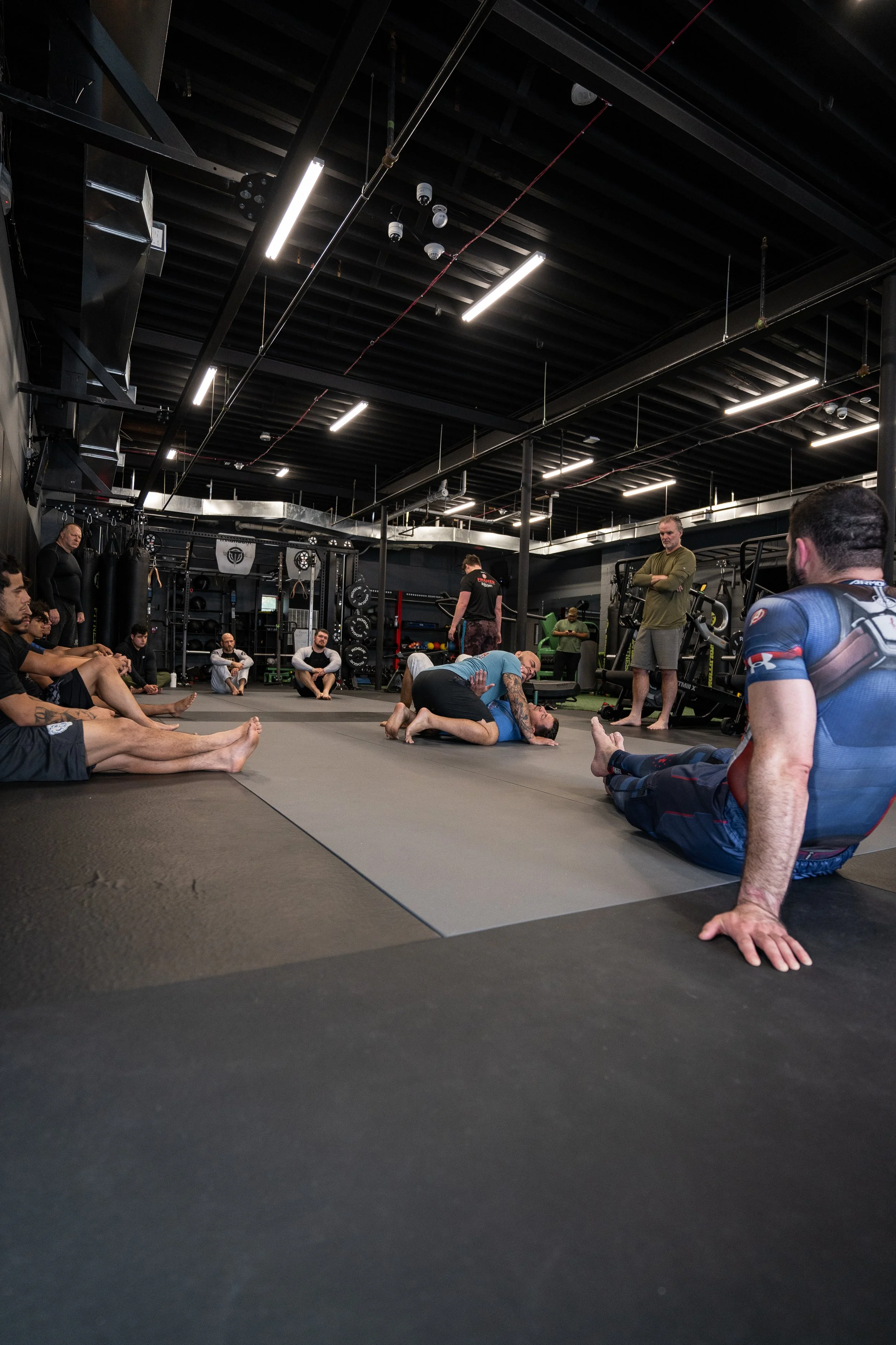 Martial arts training session in a gym with participants practicing grappling techniques on mats, surrounded by gym equipment and instructors observing.