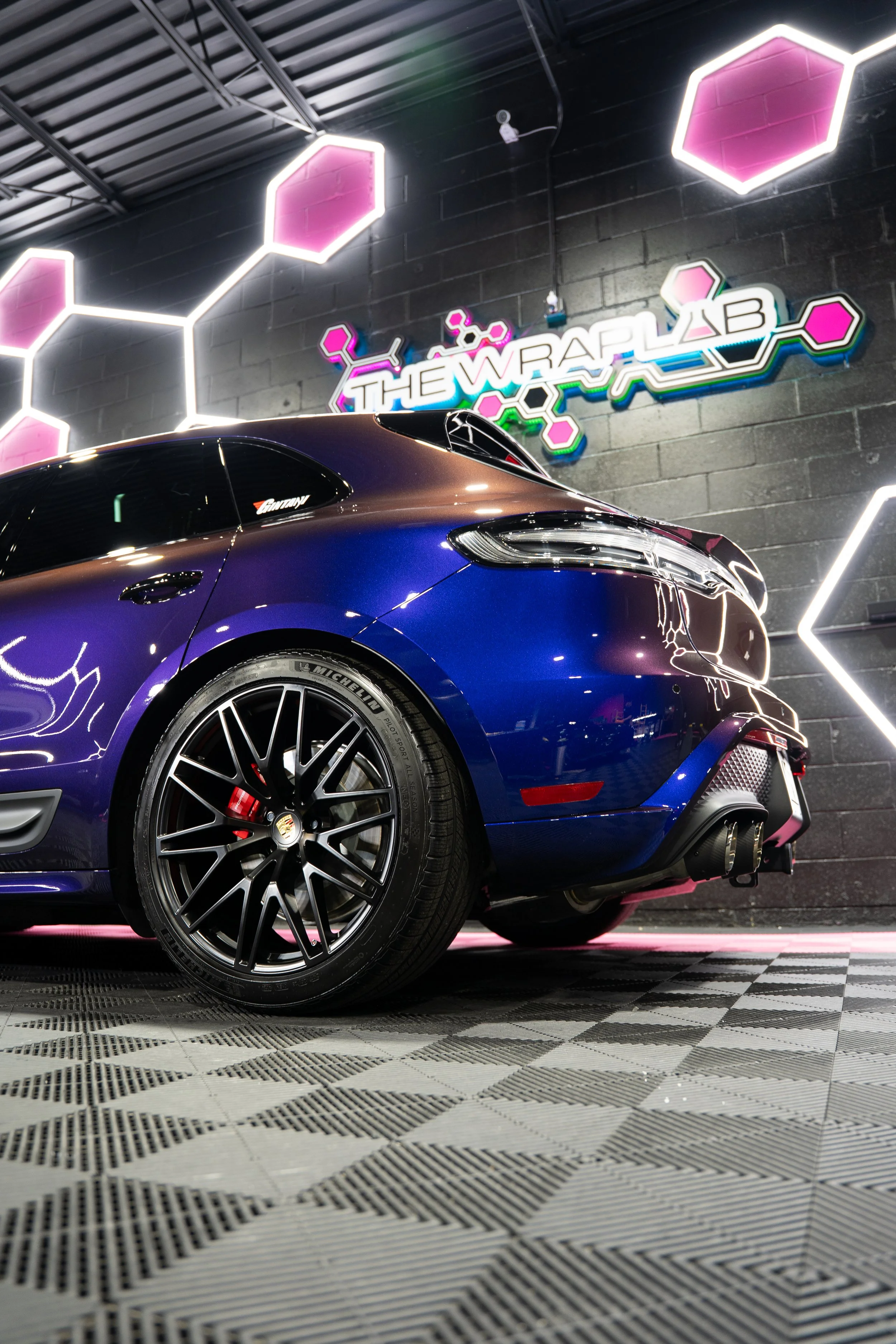A blue sports car with black wheels and red brake calipers parked indoors at a neon-lit garage with a wall sign that reads 'The Wrap Lab' and pink hexagon-shaped lights.