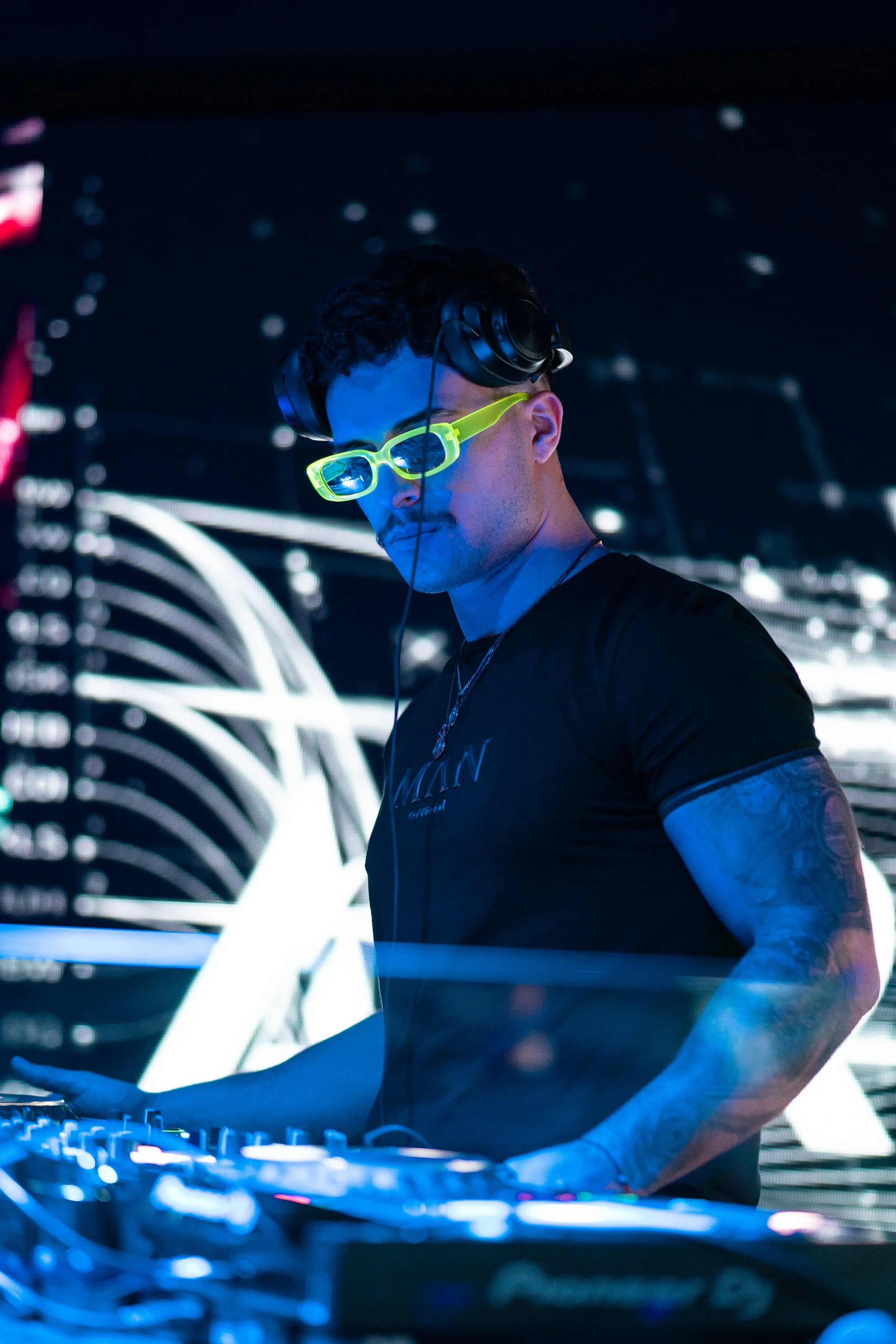 A DJ wearing black headphones and neon green sunglasses, focused on mixing music at a DJ booth with digital equipment under blue and white lighting.